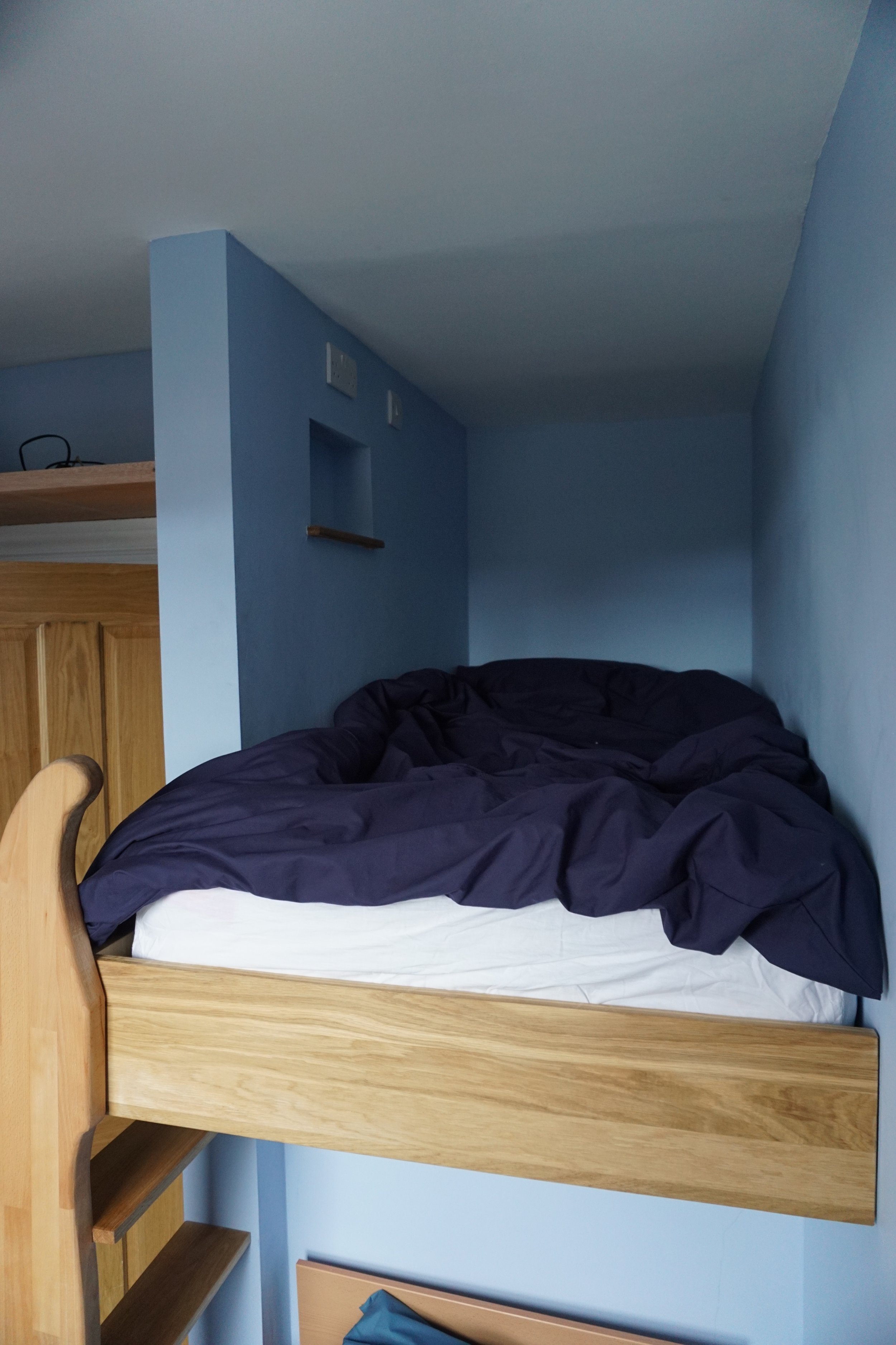 Loft bed with a black blanket and white mattress in a small blue-walled room.