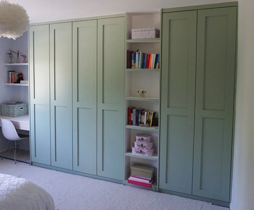 Green wardrobe with open white shelves and a small writing desk with white chair in a bedroom.