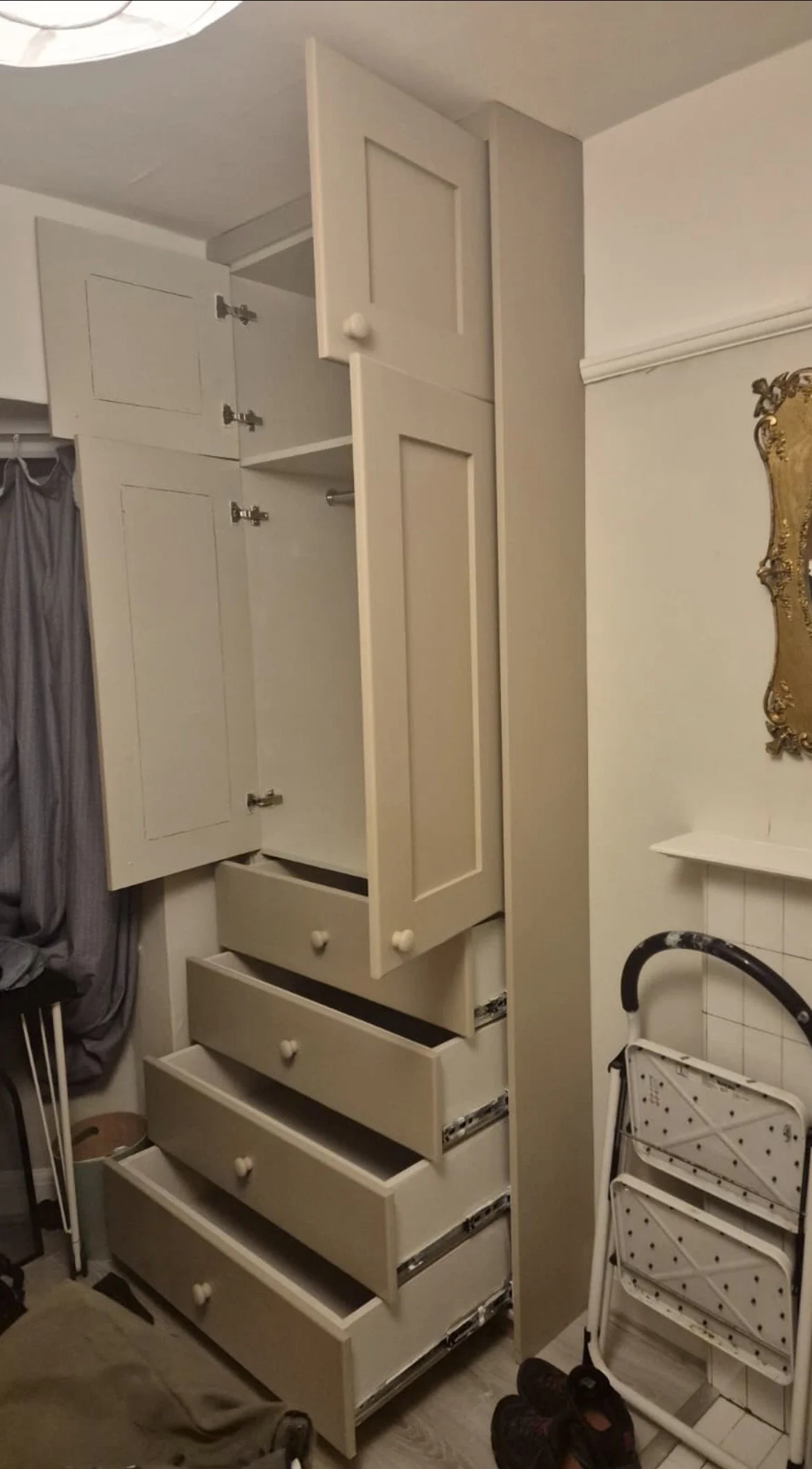 A tall, off-white wooden cabinet with open doors and six drawers below, situated in a room with gray curtains on the left and a white wall with ornate gold-framed mirror on the right. Shoes and a folding drying rack are on the floor.