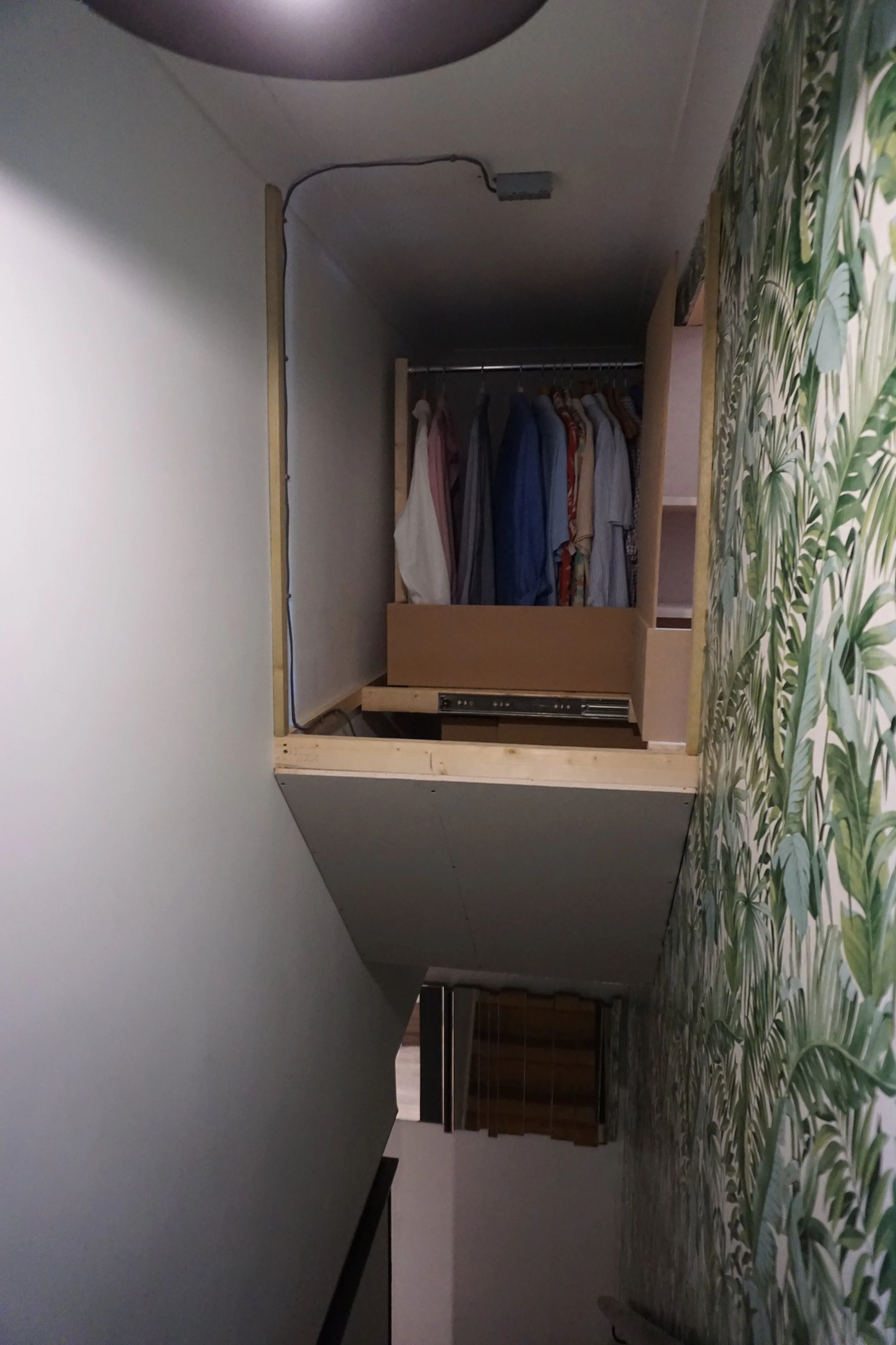 A small, unfinished closet with hanging clothes and wooden shelving, stairs visible in the foreground, and a wallpapered wall with a leaf pattern on the right side.