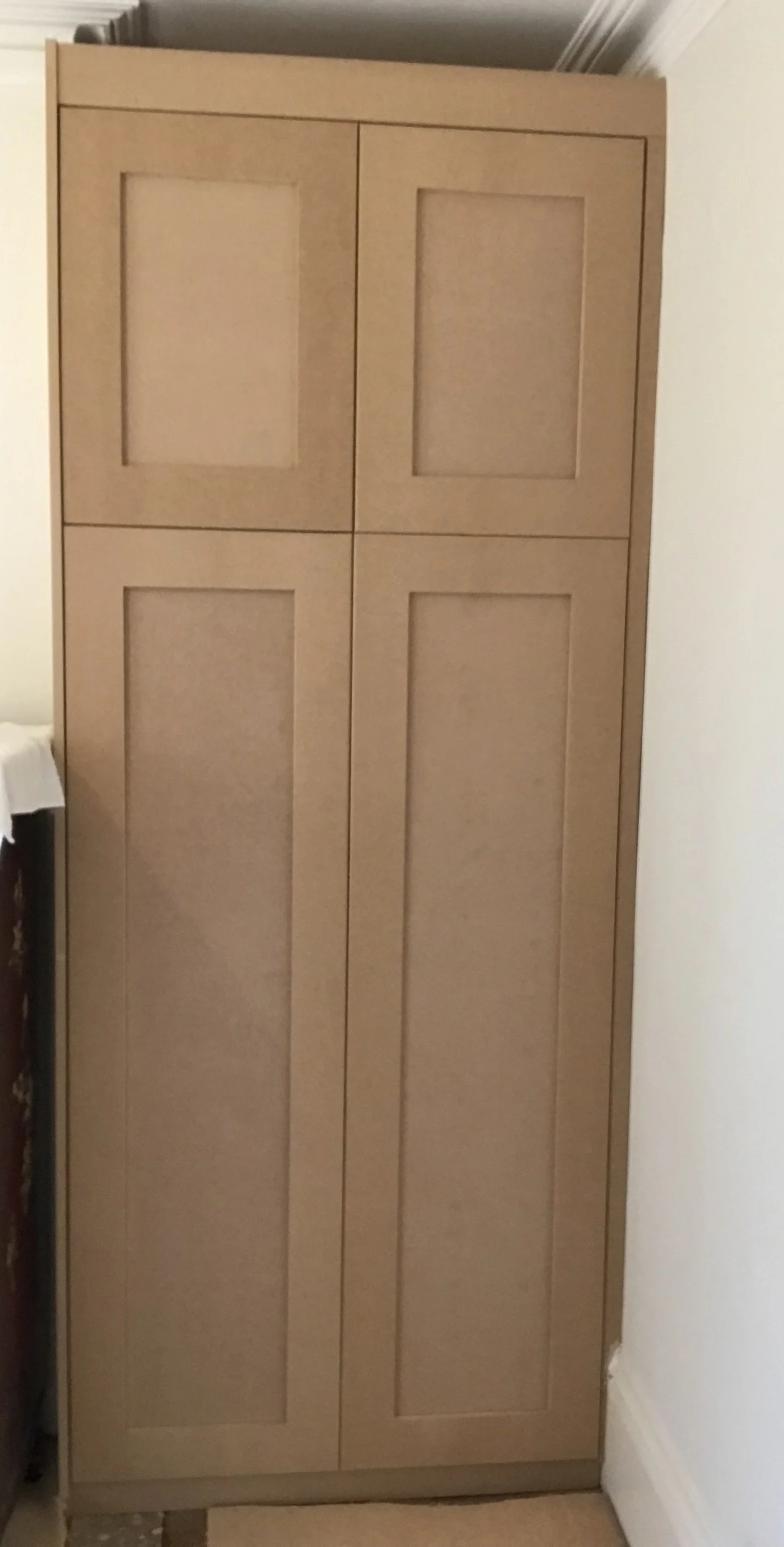 A wooden cabinet with four rectangular panels and a natural wood finish standing against a white wall.