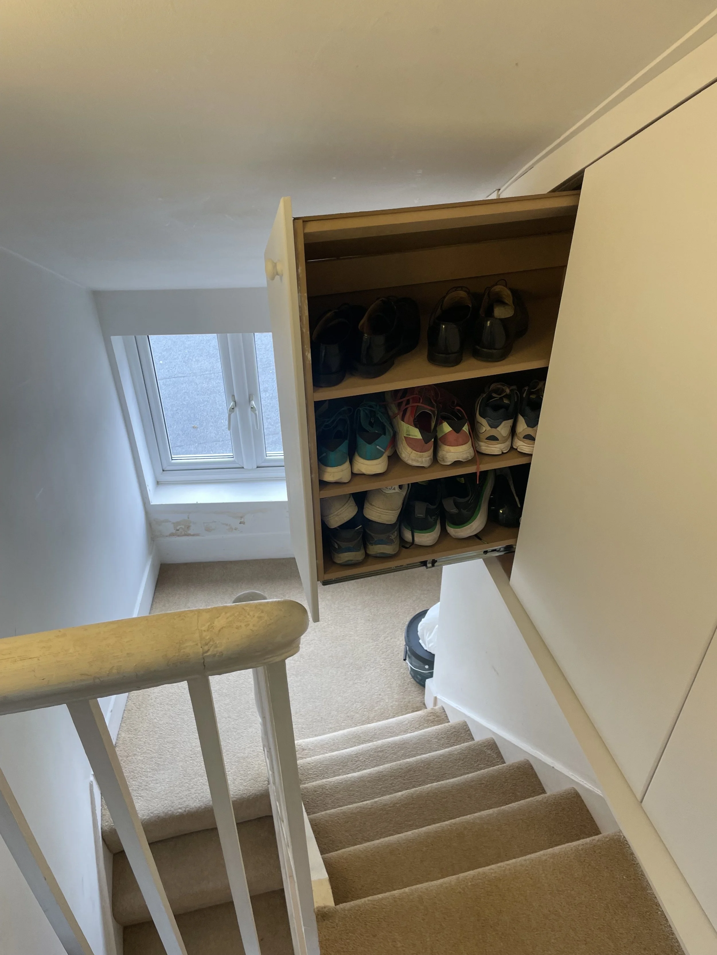 A view from the top of a staircase showing a hallway with a built-in shoe cabinet filled with various shoes next to a window.