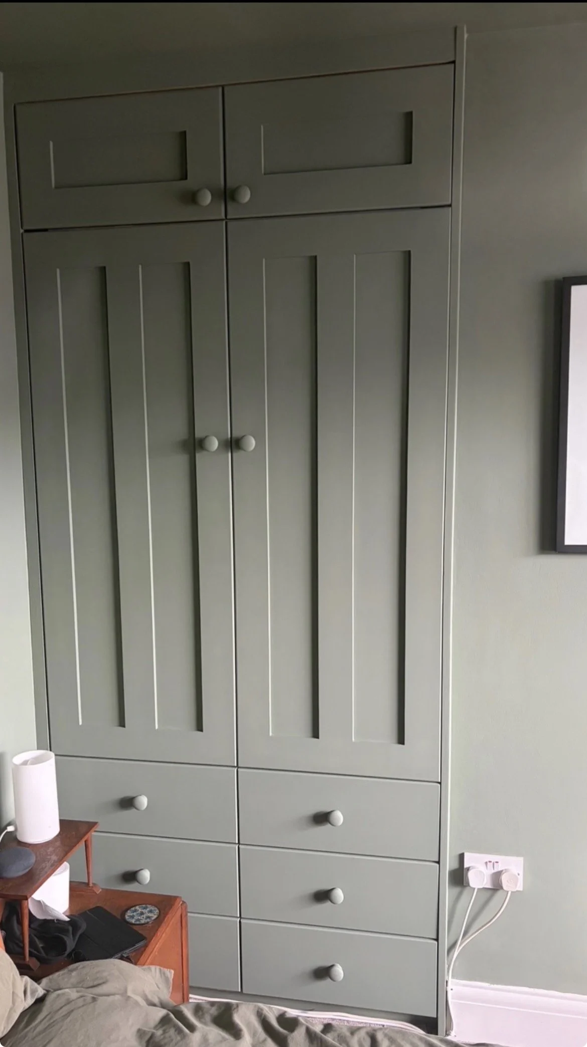 A tall, gray cabinet with panel doors and round knobs in a room with a green wall. A small bedside table with decor items is partially visible in the foreground.