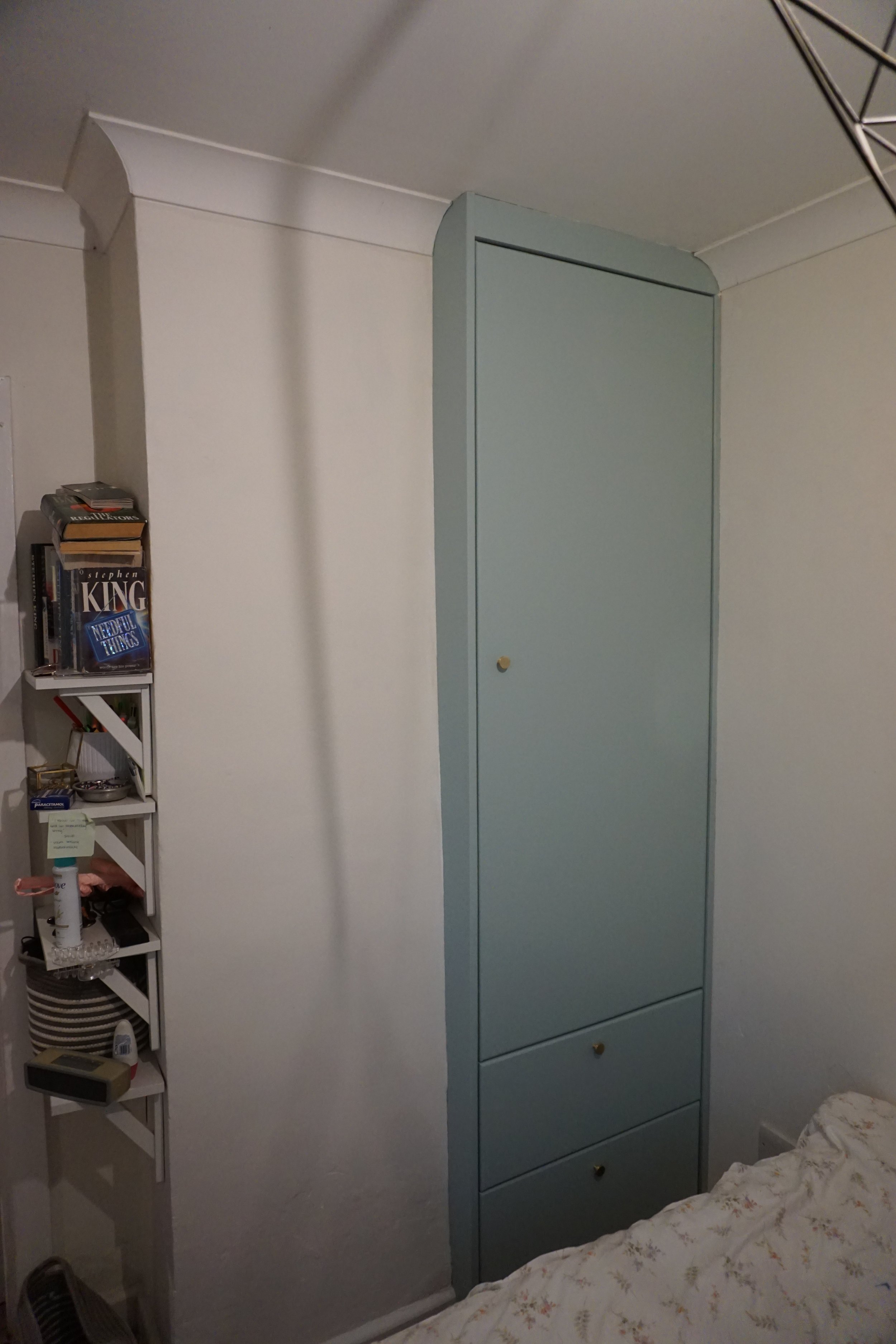 A small bookshelf filled with books, medications, and miscellaneous items stands next to a built-in wardrobe with a rounded top and three drawers underneath, in a bedroom with white walls.