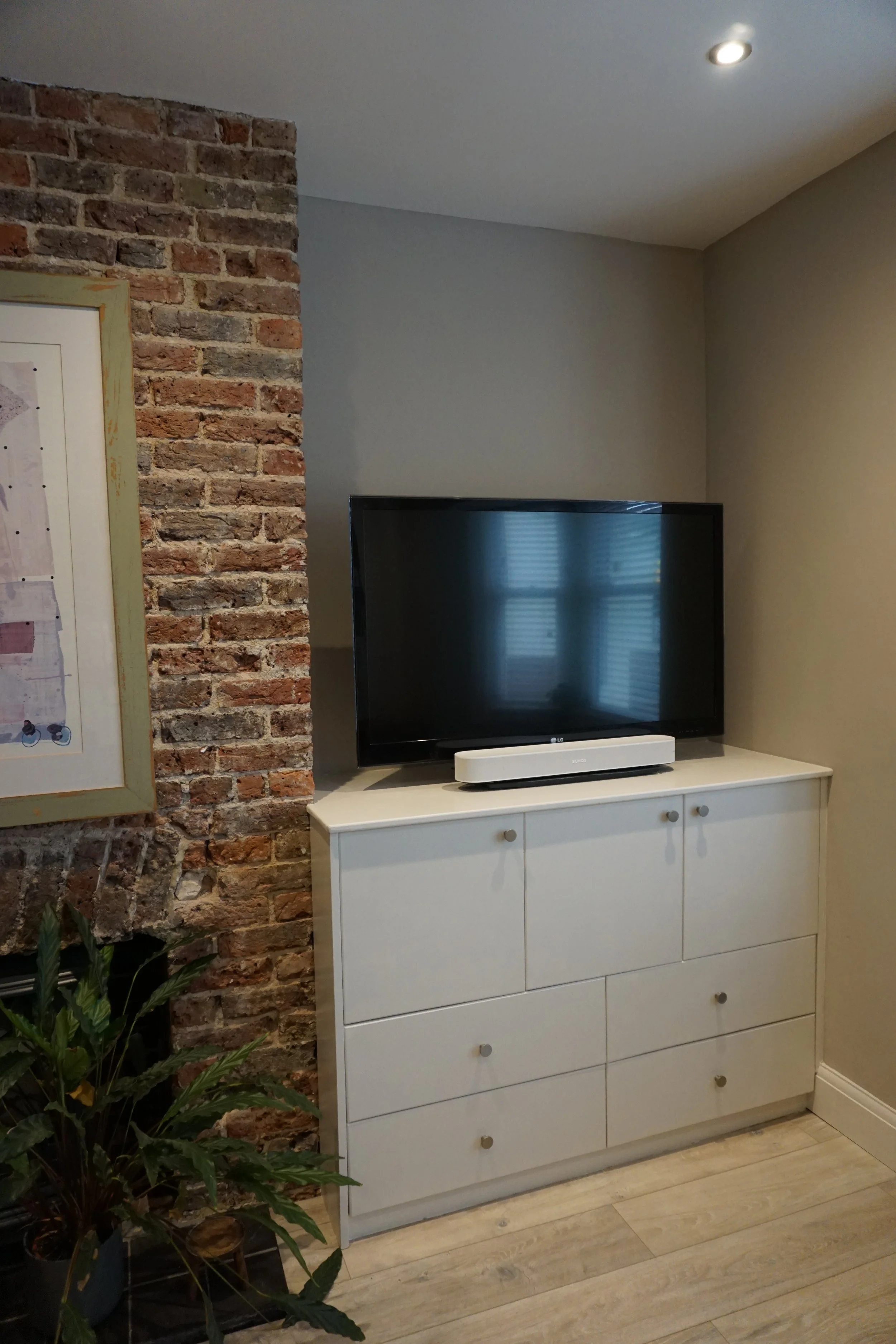 A living room corner with a flat-screen TV on a white cabinet, a brick wall on the left, and a potted plant in the bottom left corner.