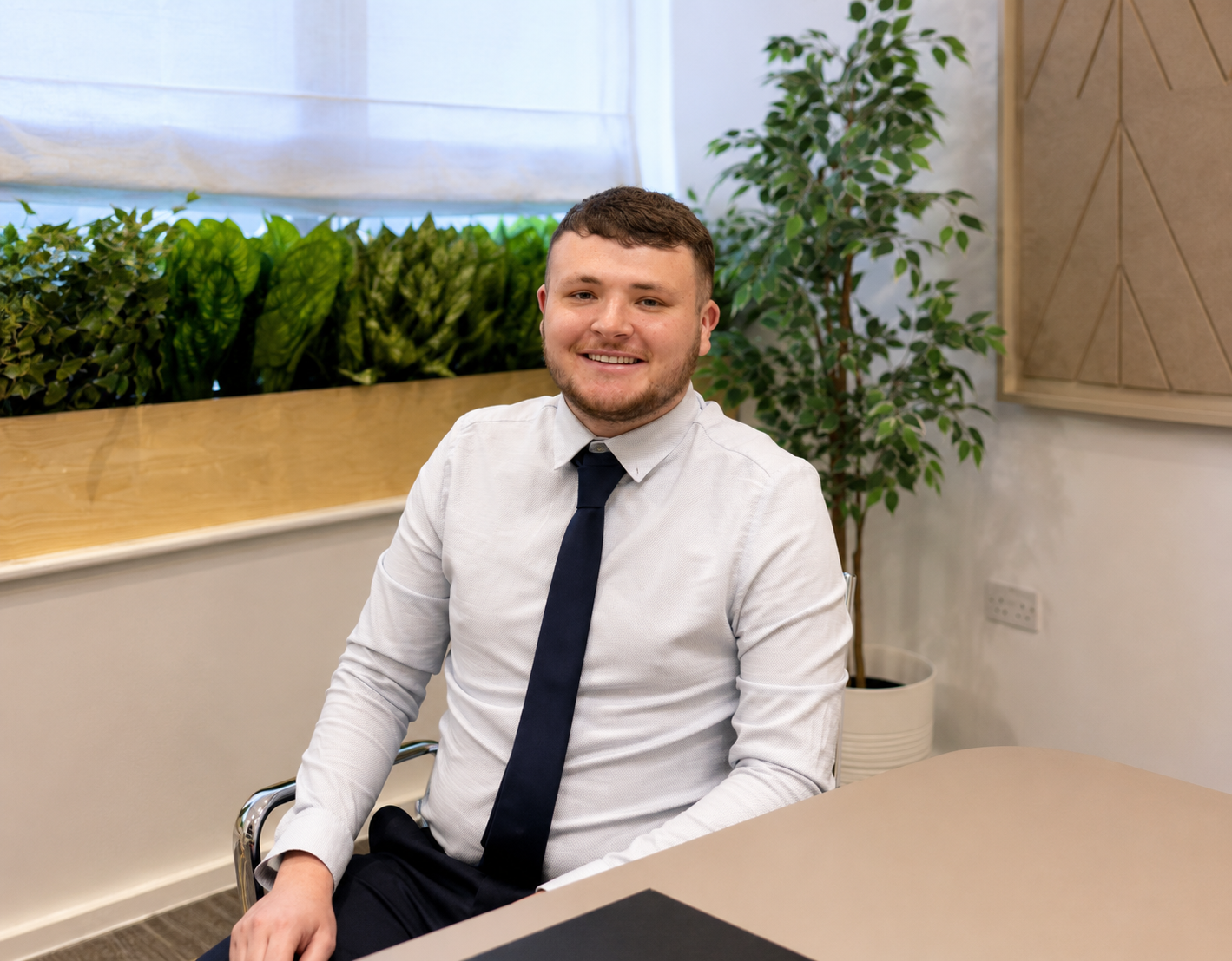 Jack Pettigrew seated in a modern office meeting room wearing a white shirt and navy tie, smiling at the camera with indoor plants in the background.
