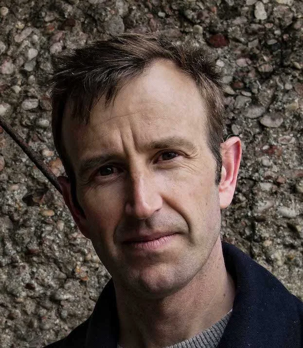 Close-up of a man with short brown hair and a slight smile, standing outdoors against a rocky background.