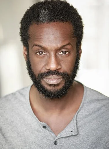 Close-up portrait of a man with dark skin, short curly hair, and a full beard, wearing a light gray shirt, looking directly at the camera.