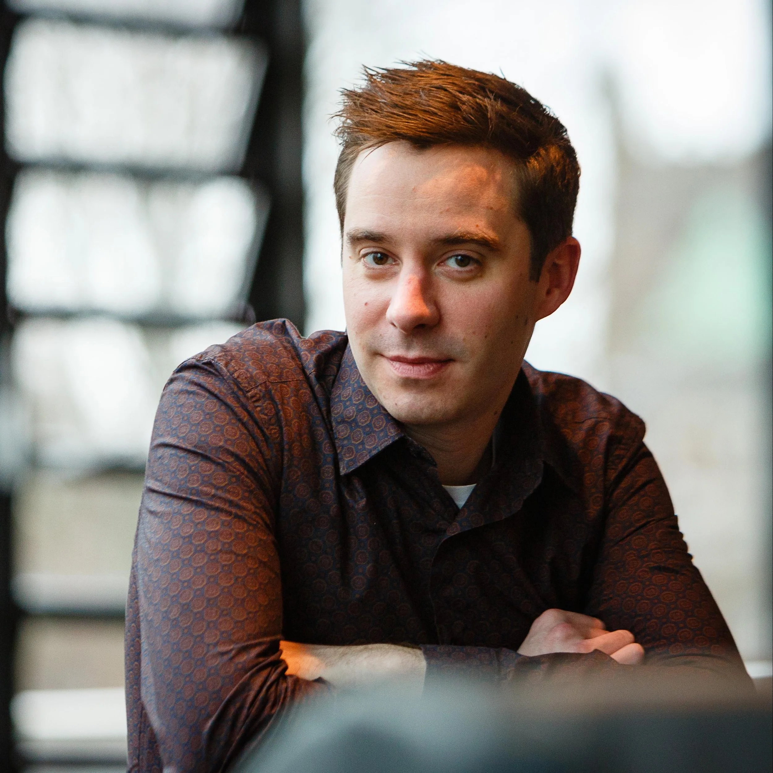 A young man with short brown hair and light skin sitting at a table, wearing a patterned shirt, indoors with blurred windows in the background.