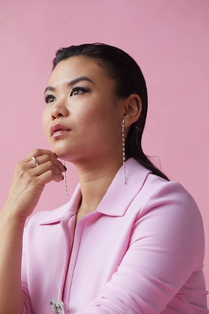 A woman with long dark hair styled back, wearing a pink blazer with a decorative pin, silver drop earrings, and a silver ring, posing against a pink background.