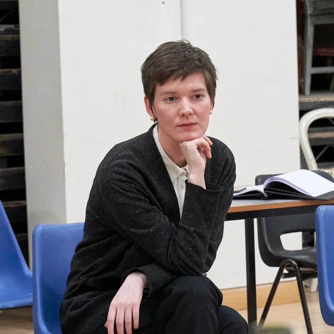 A young woman with short brown hair wearing a black speckled blazer and white shirt, sitting at a table with an open notebook, in a classroom or meeting room.