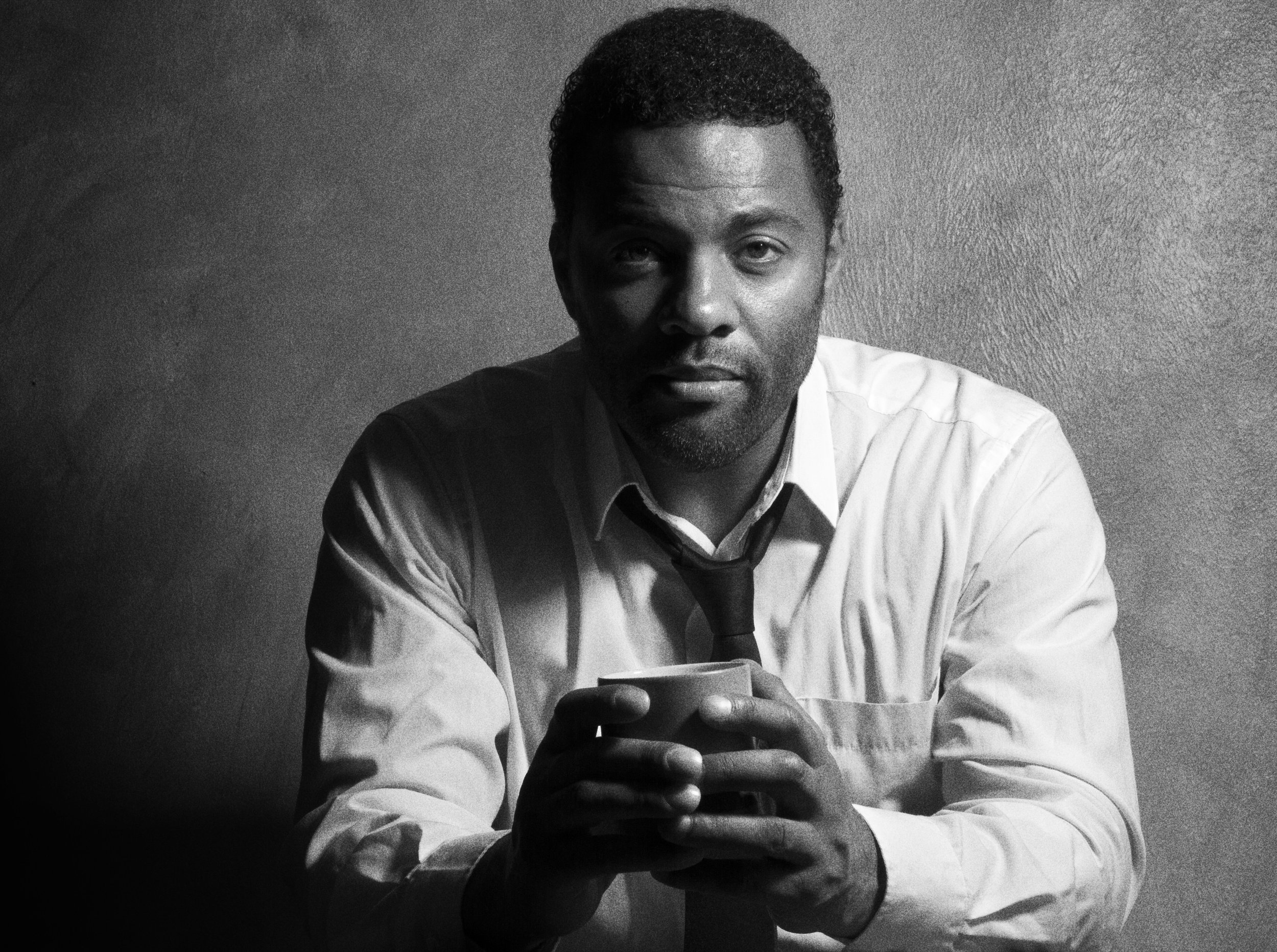 A black man with short hair and a classic white shirt and black tie. He's holding a cup and looking straight into the lens.