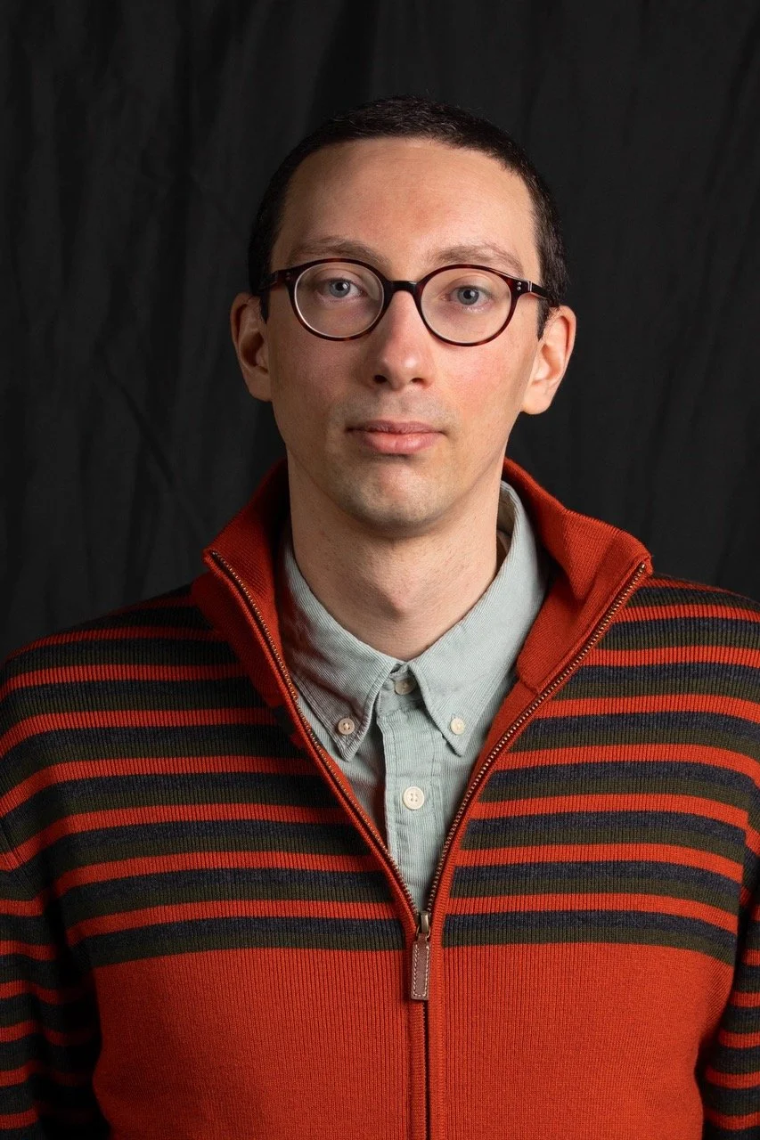 Portrait of a man with glasses wearing a striped red and black sweater over a light gray button-up shirt, against a dark background.