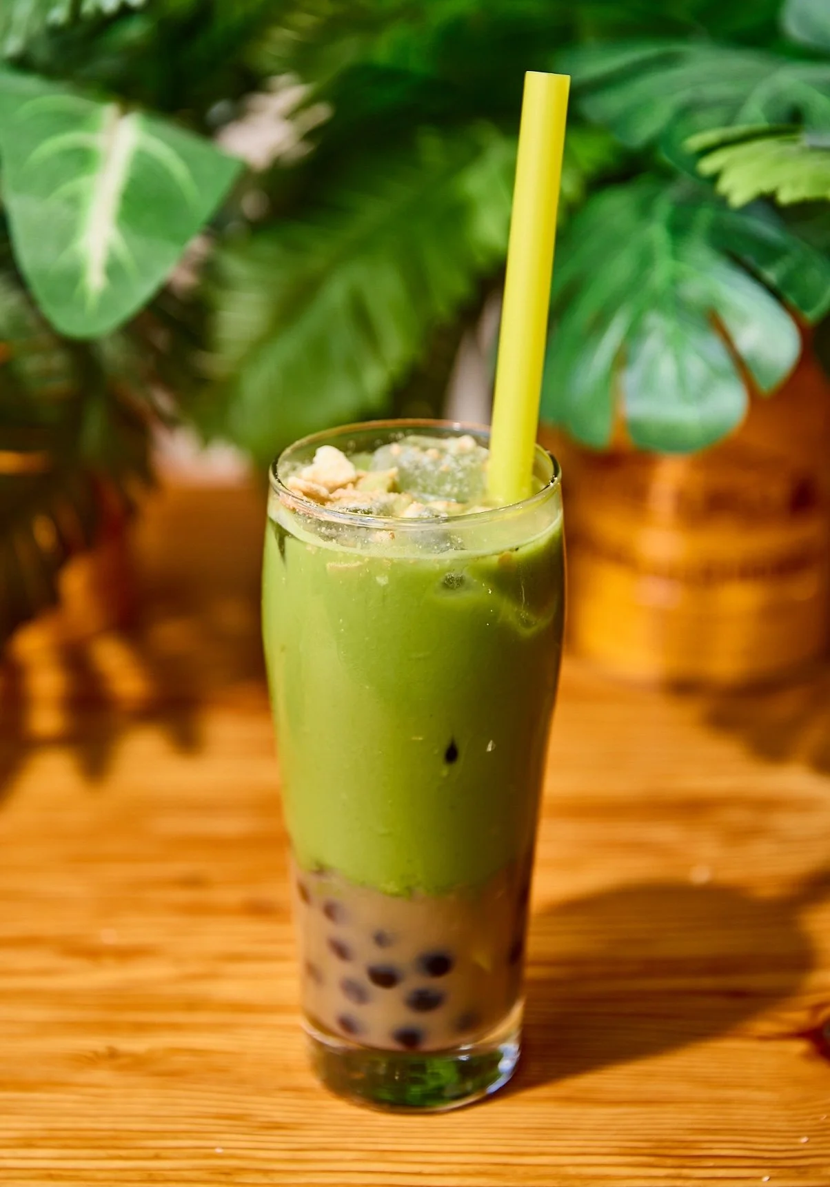 A tall glass of green matcha bubble tea with black tapioca pearls at the bottom and a yellow straw on a wooden table, with green leafy plants in the background.