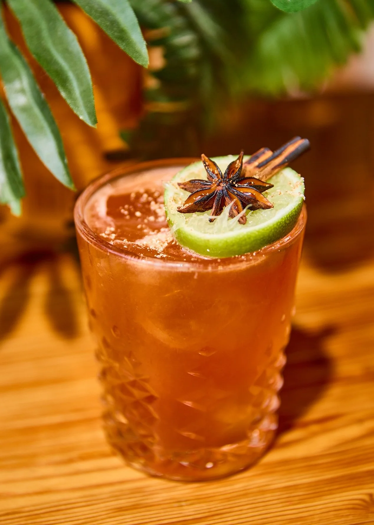 A cocktail garnished with a slice of lime, star anise, and cinnamon sticks on a wooden surface.