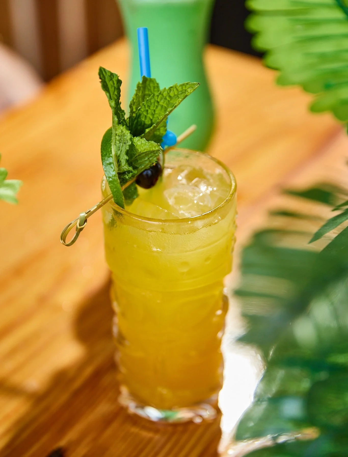 A tall glass of yellow cocktail with ice, garnished with mint, lime, and a cherry, with two blue straws, placed on a wooden table, surrounded by green plants.