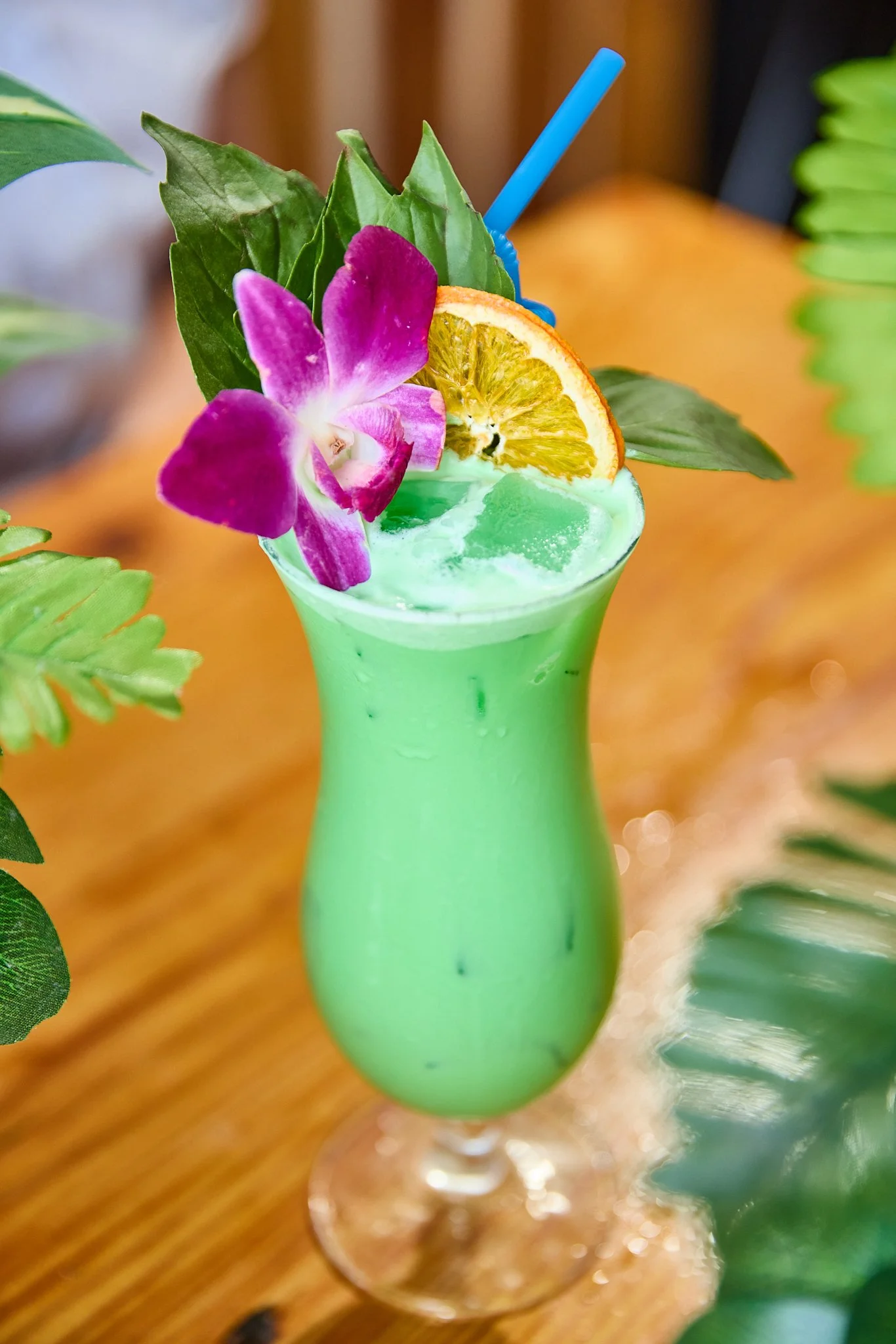 Green tropical cocktail topped with an orange slice, purple orchid, greenery, and a blue straw, placed on a wooden surface surrounded by green leaves.