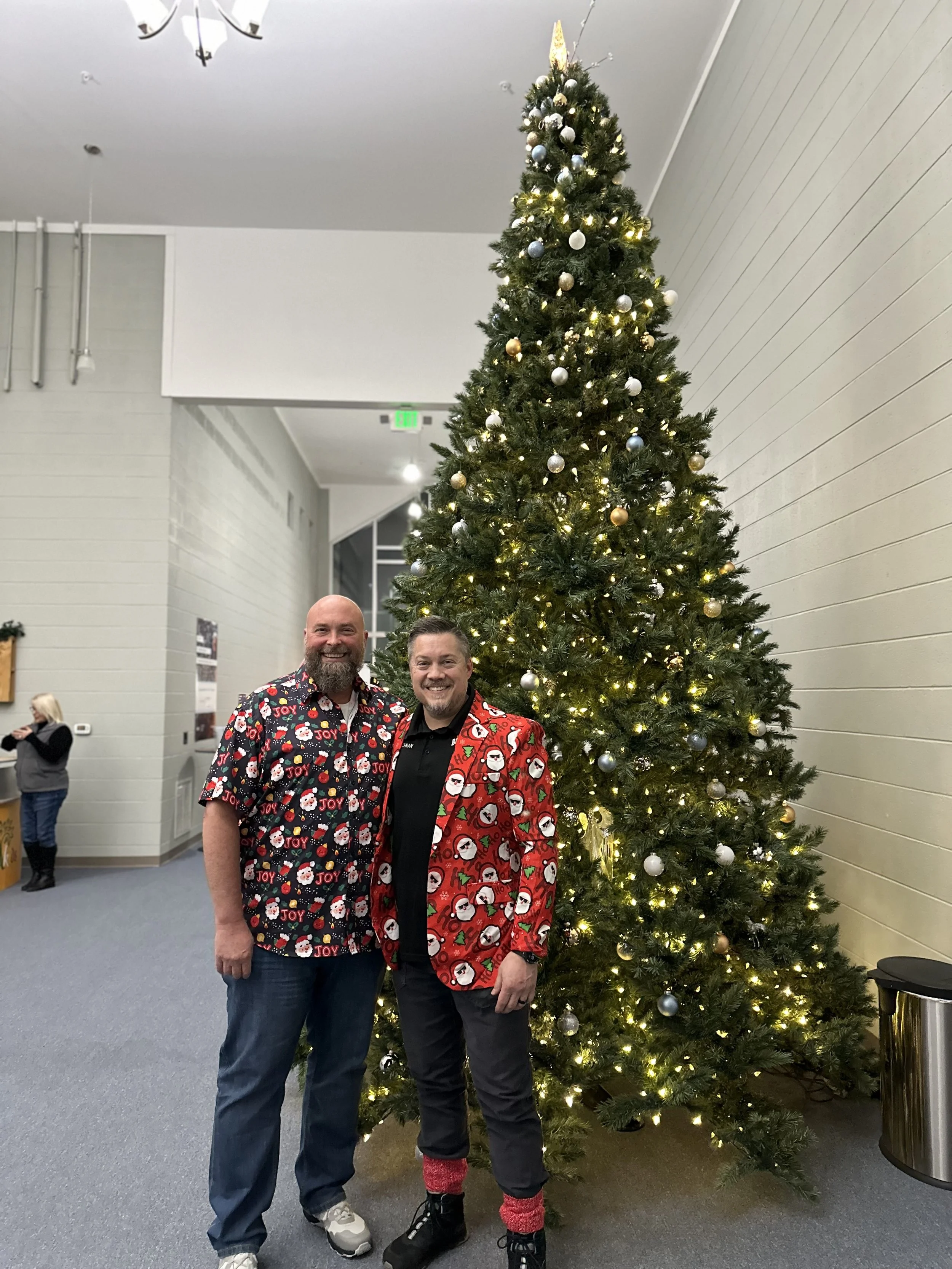Two men dressed in festive Christmas shirts stand in front of a decorated Christmas tree with lights and ornaments, smiling at the camera.