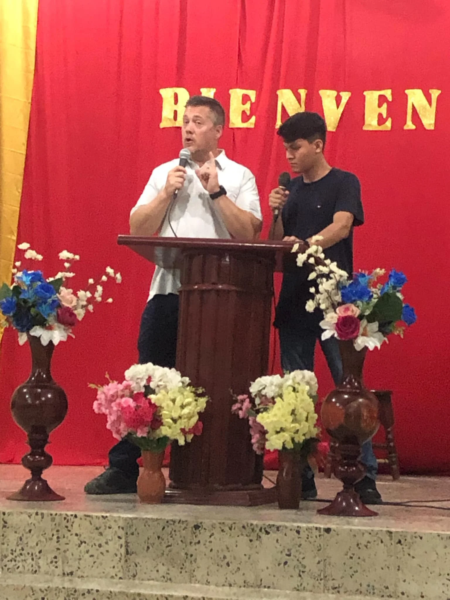 Two men standing behind a wooden podium with microphones, a man in white shirt speaking and gesturing with his index finger, a young man in black shirt holding a microphone, floral arrangements on the stage, red curtain backdrop with gold letters spe