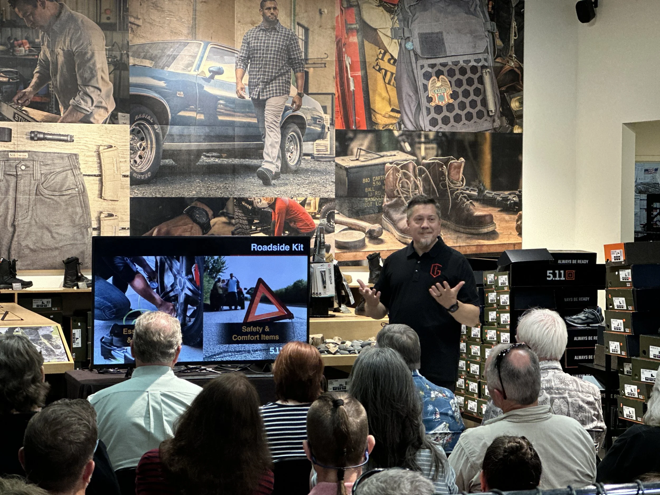 A man giving a presentation to an audience in a retail store, with a display of outdoor gear, shoes, and a large screen showing slides about roadside kit and safety items.