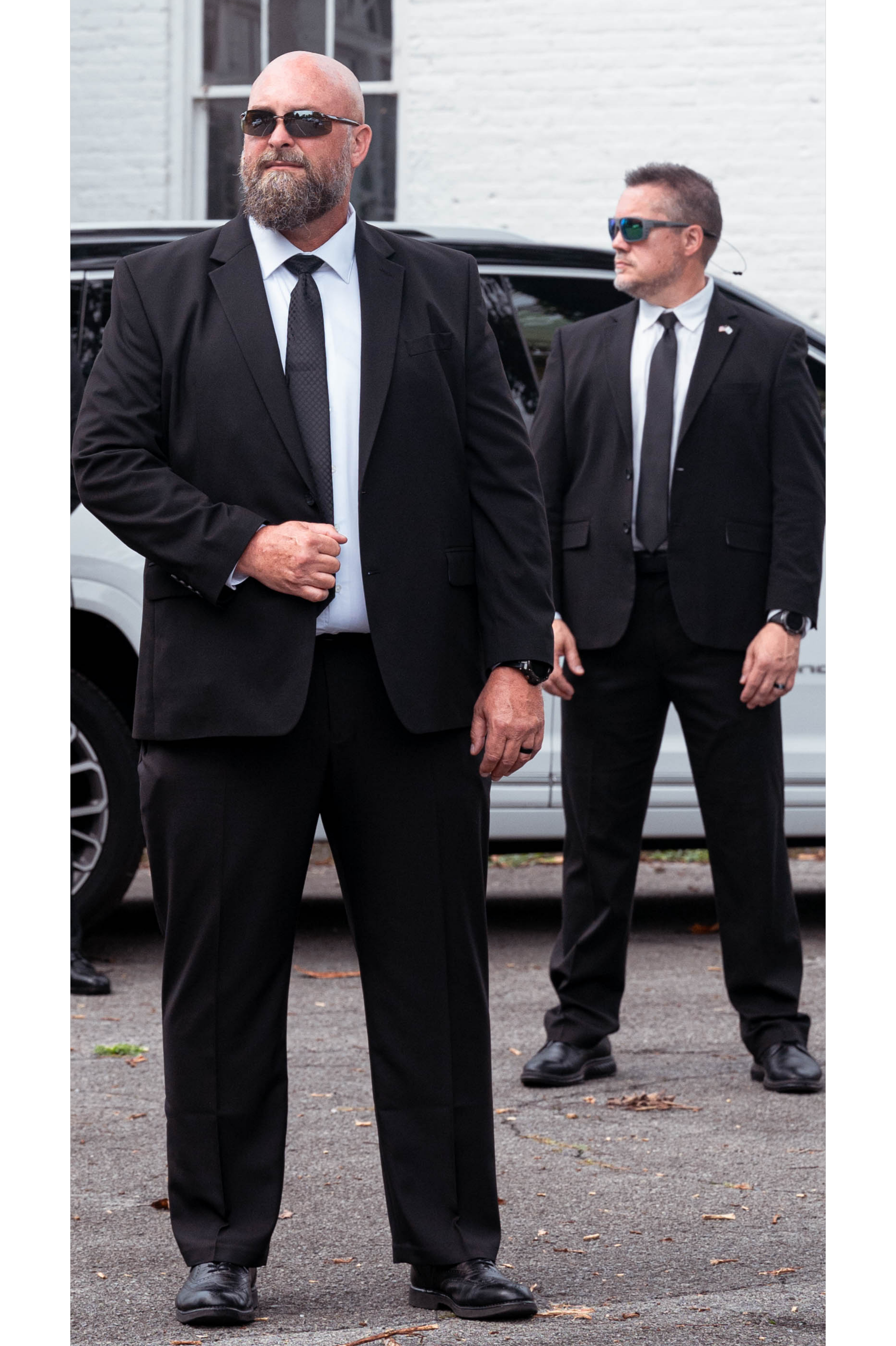 Two men in black suits and sunglasses standing outside near a vehicle, with a white brick building in the background.