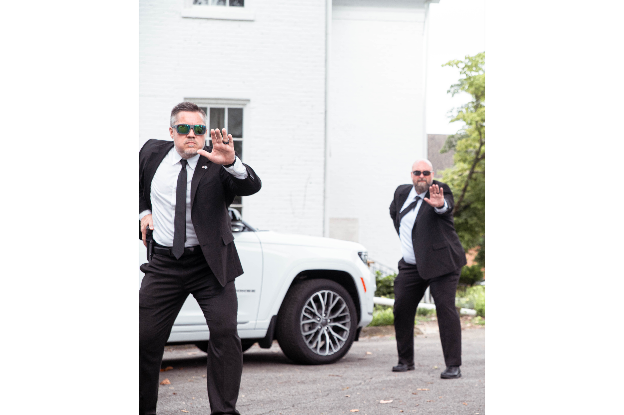 Two men dressed like secret service agents in suits, sunglasses, and black ties, standing in front of a white vehicle and a white building, holding up their hands in a stop gesture.