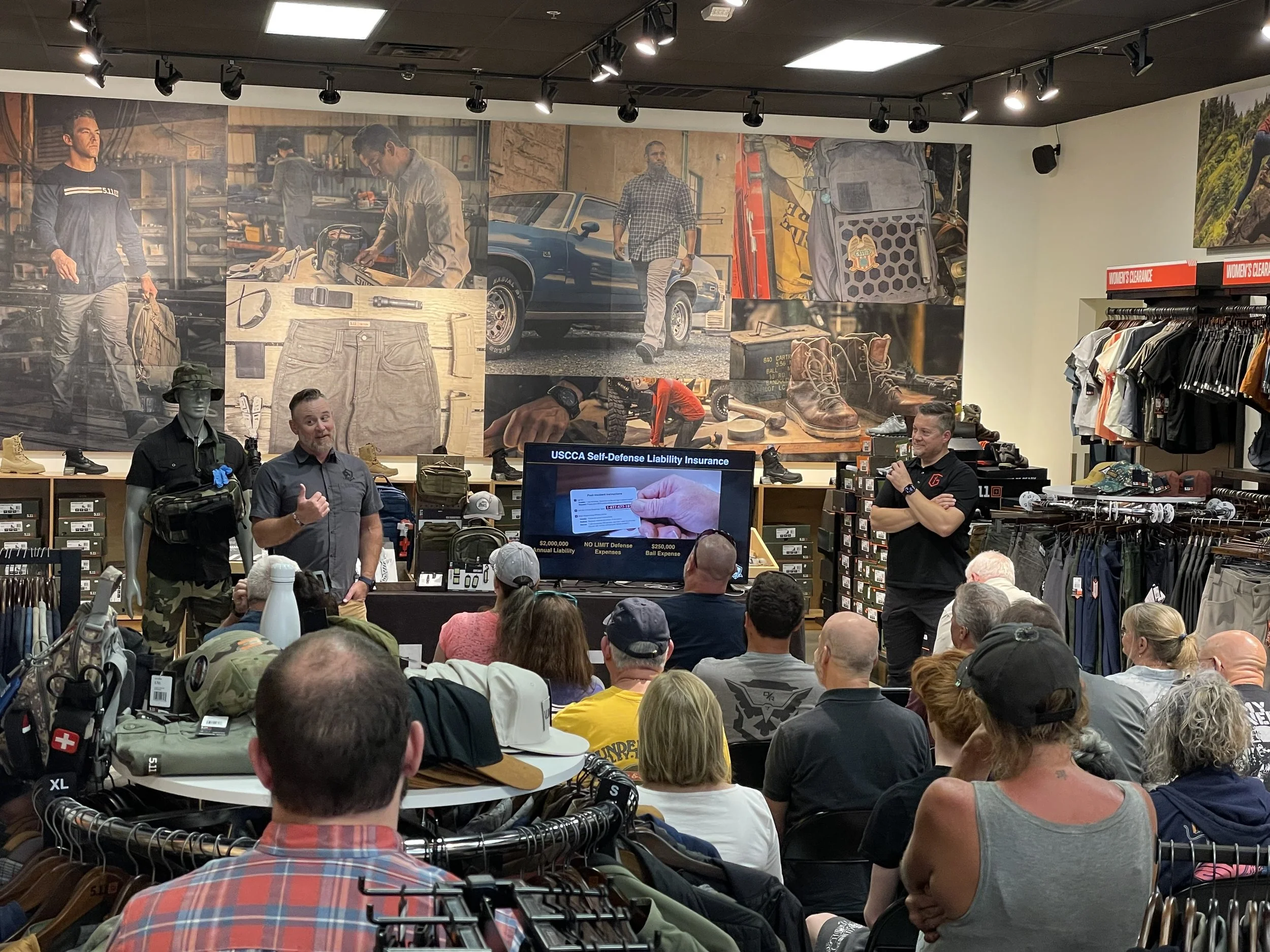 Man giving a presentation to an audience in a retail store, with a screen displaying 'USCCA Self-Defense Liability Insurance' and various outdoor gear and clothing items surrounding them.