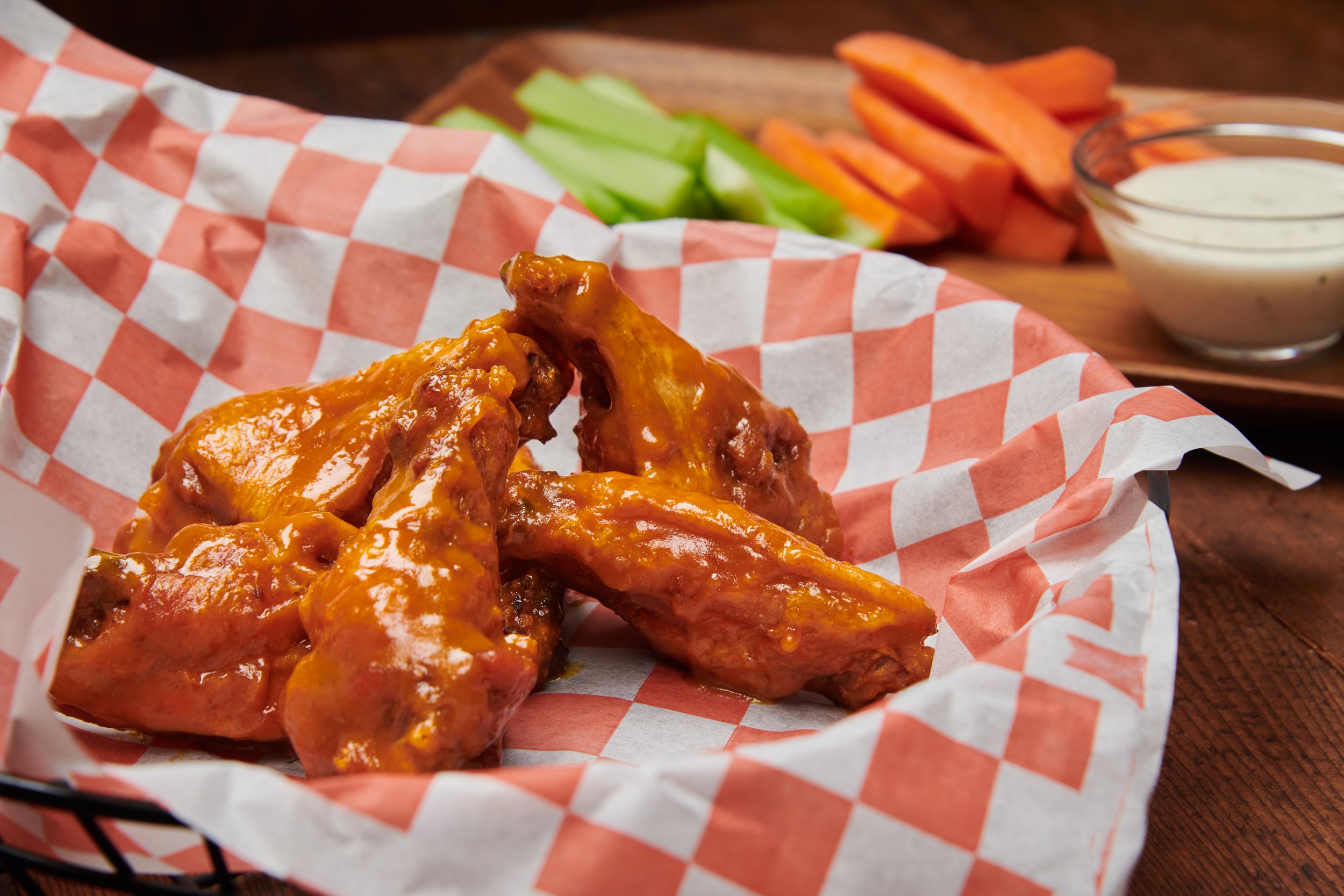 Chicken wings in a basket lined with red checkered paper, with celery sticks, carrot sticks, and a small bowl of ranch dipping sauce in the background.