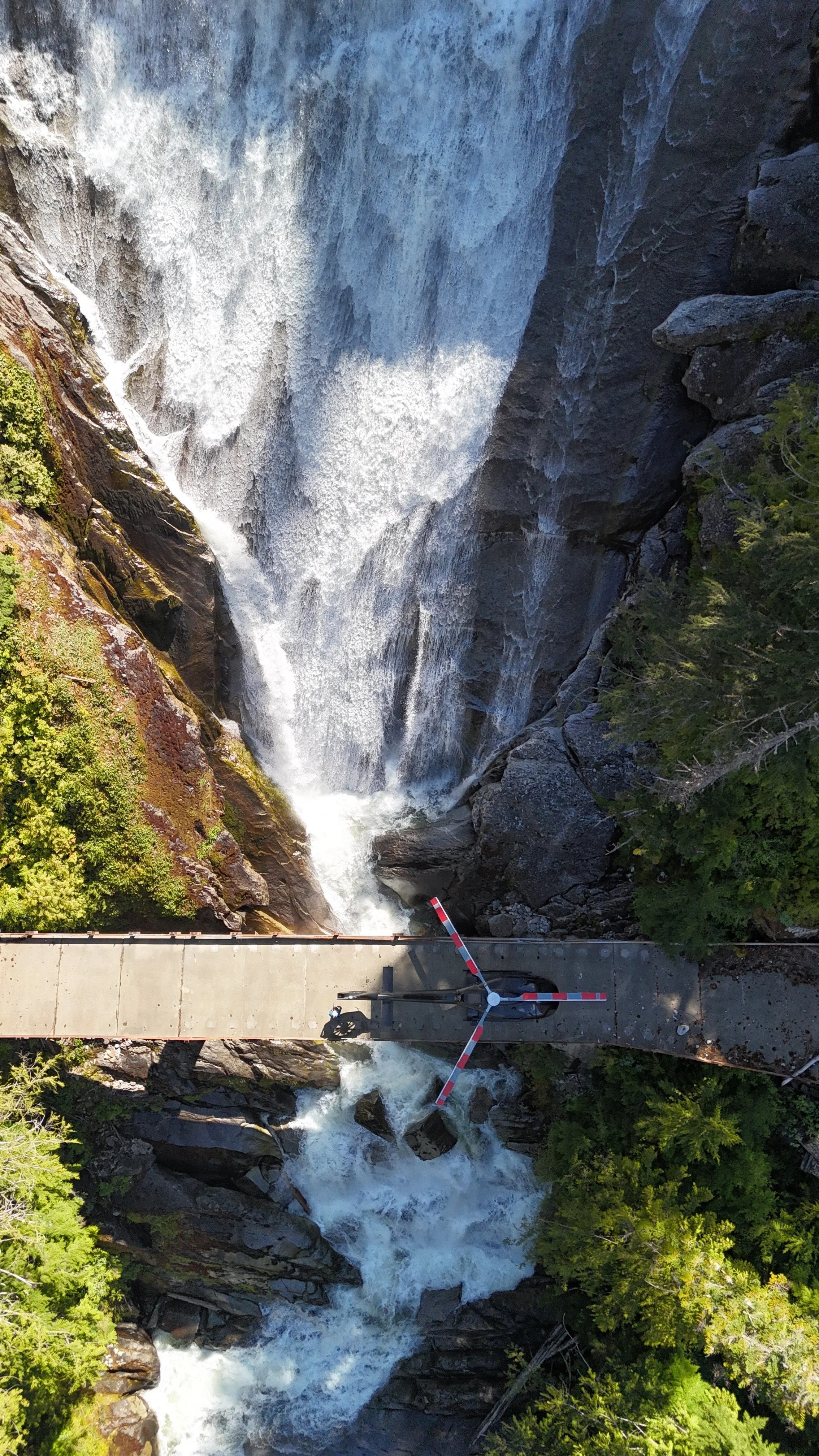 An aerial view of a waterfall next to a bridge with a red and white barrier, surrounded by green trees.