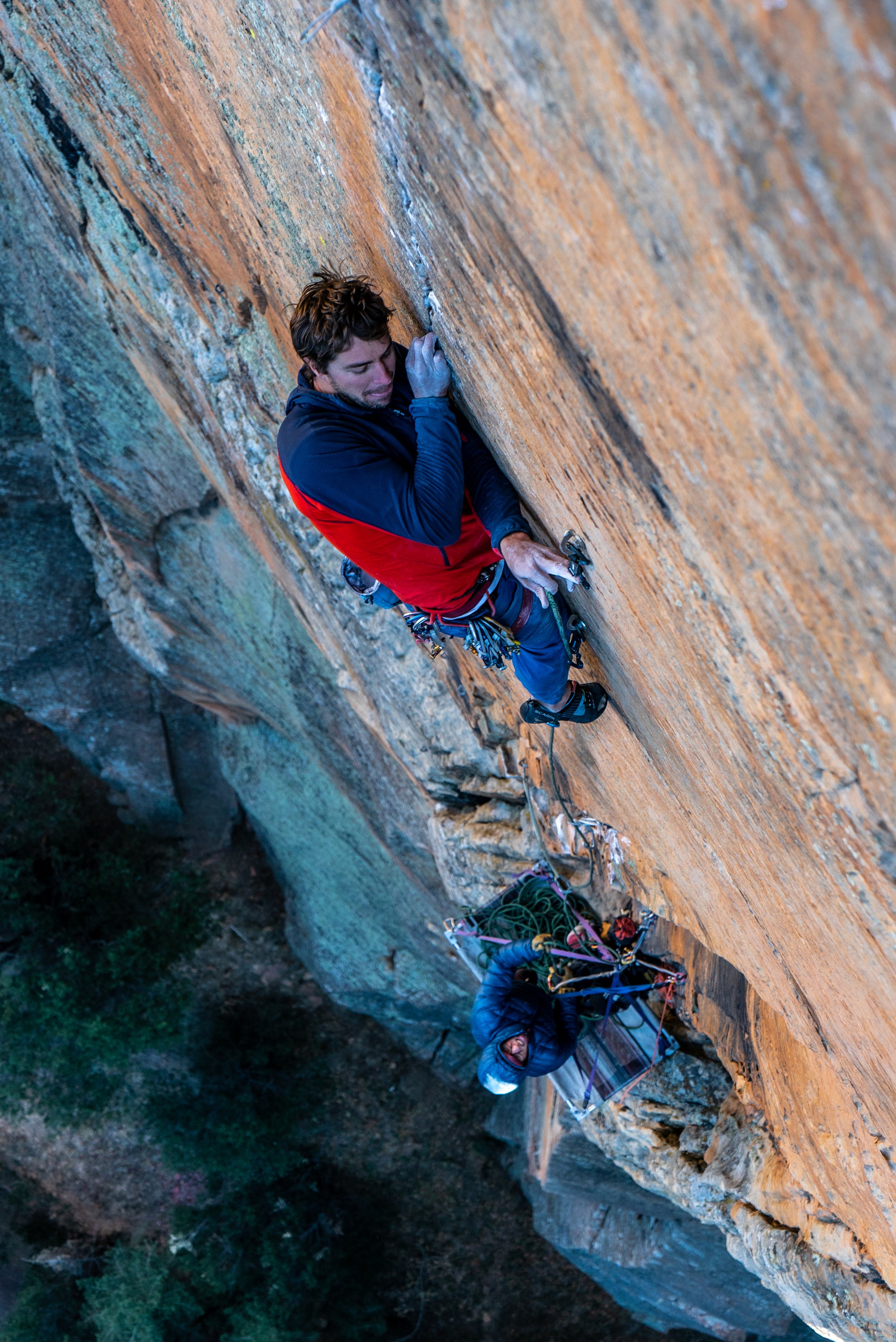 A male rock climber scaling a steep, orange sandstone cliff with safety gear, while another person at the base manages climbing equipment.