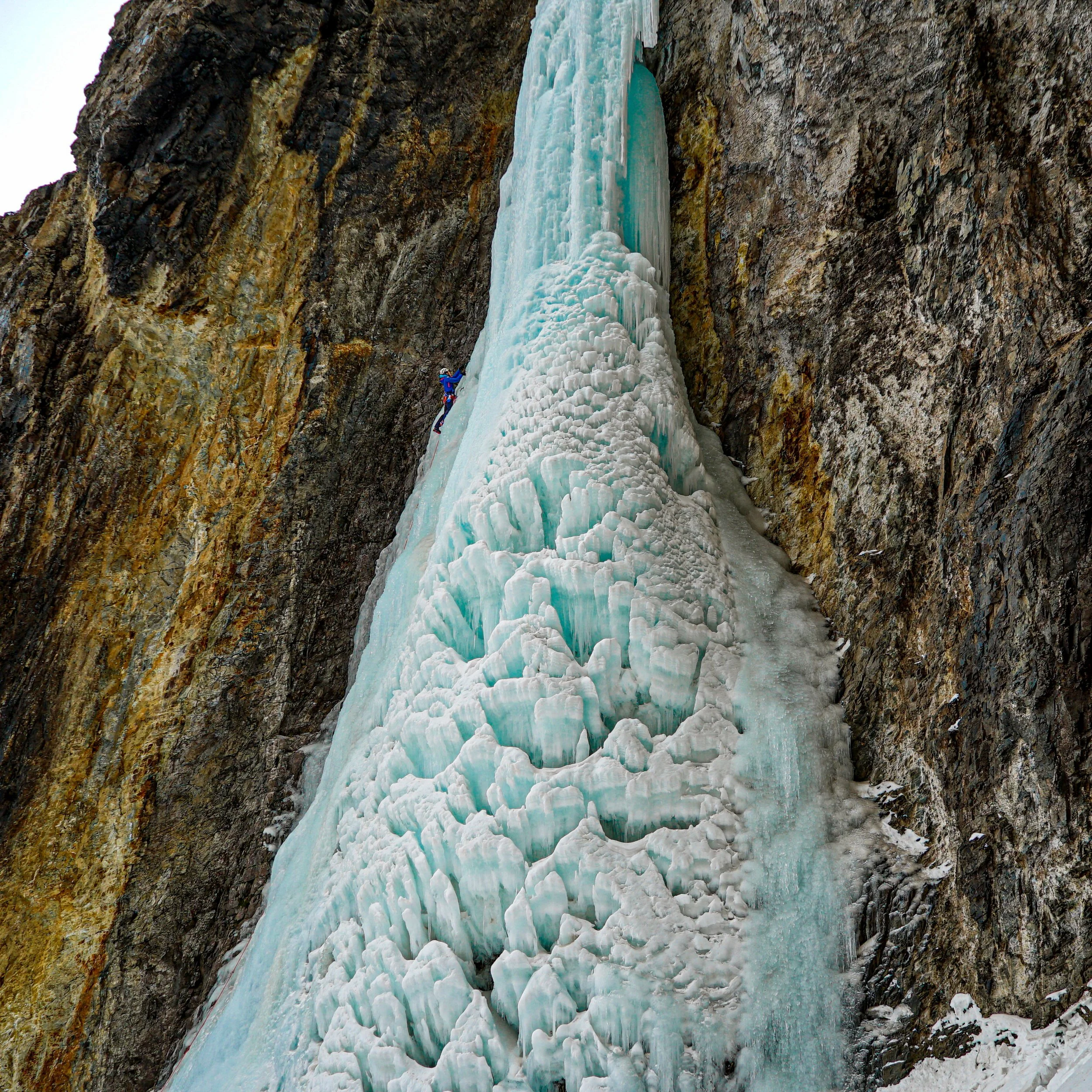 Ice climber ascending a frozen waterfall on steep rock face.