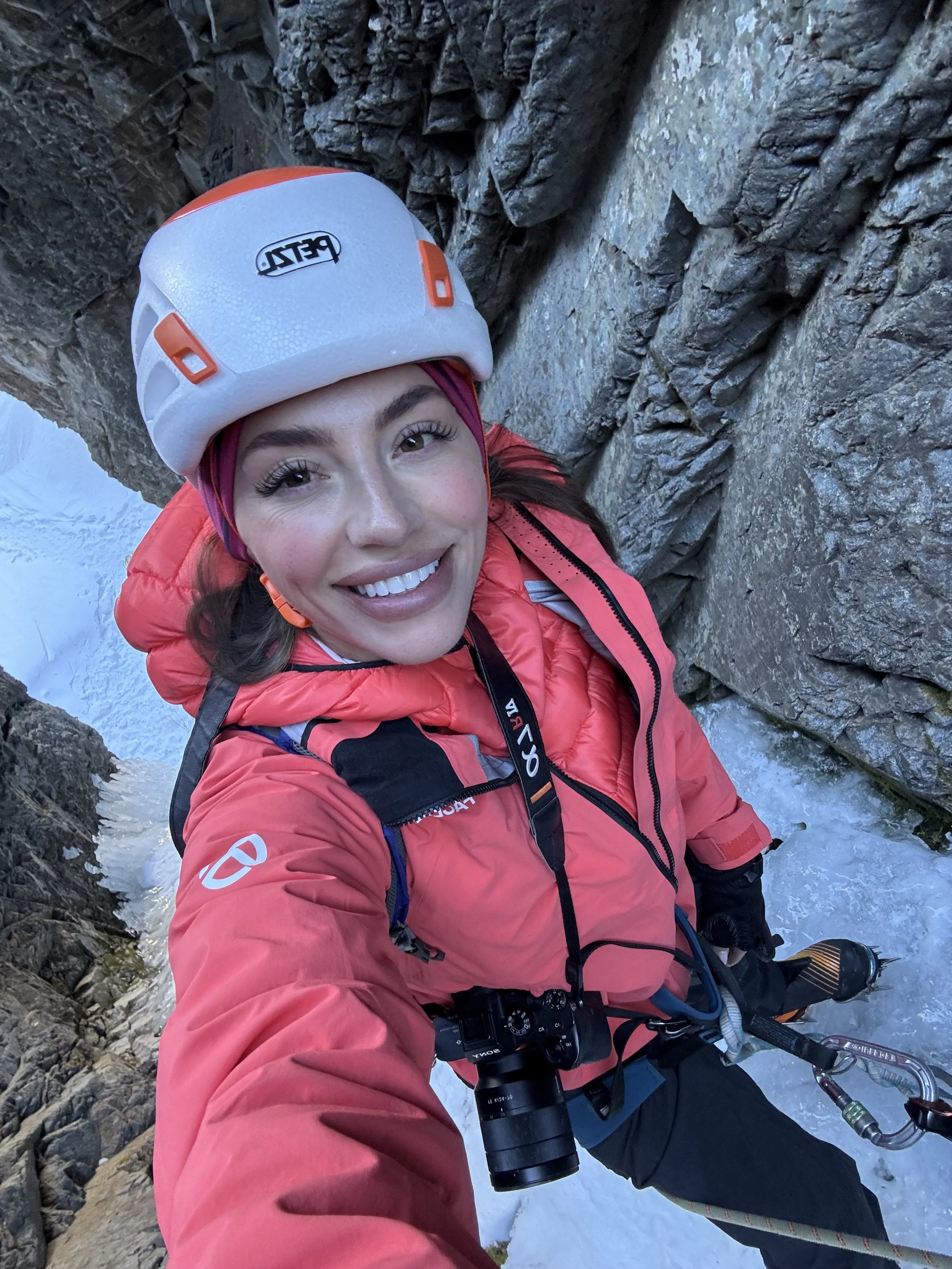 A woman in a red jacket, helmet, and gloves taking a selfie with a harness and camera, standing on a narrow snowy mountain ledge with rocky cliffs.