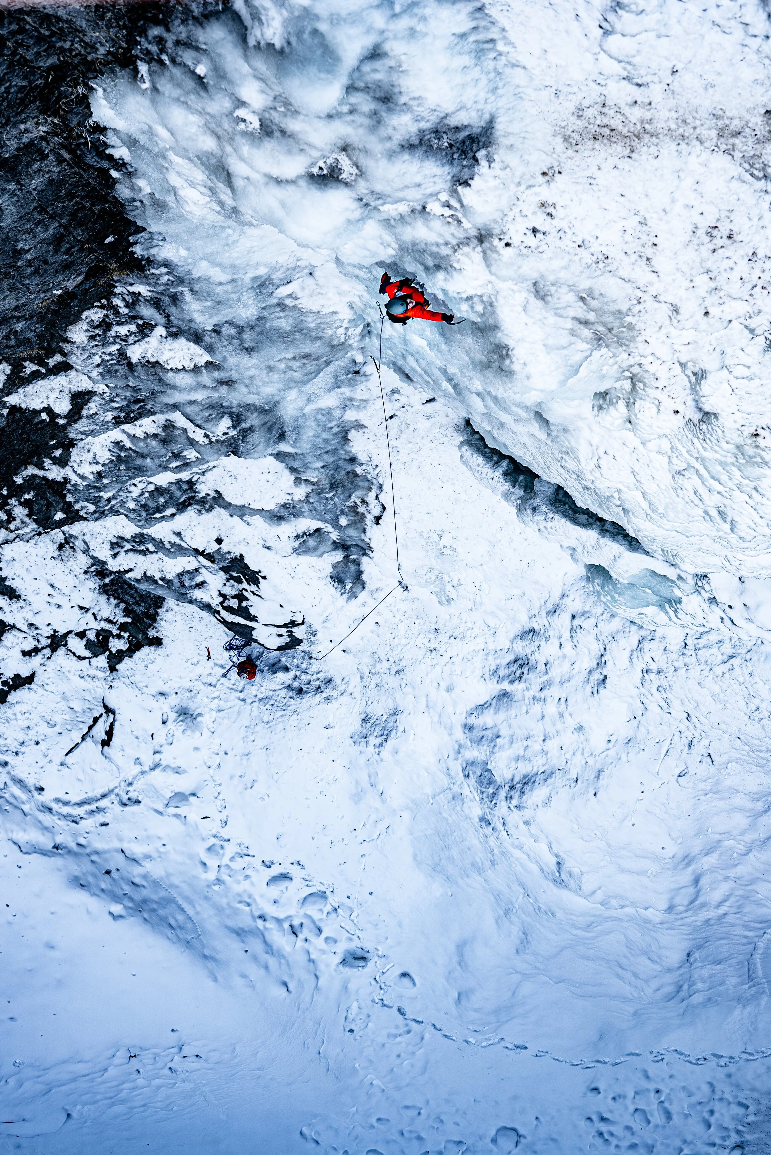 An ice climber in red gear ascending a frozen waterfall with a second climber below, on a snowy mountain.