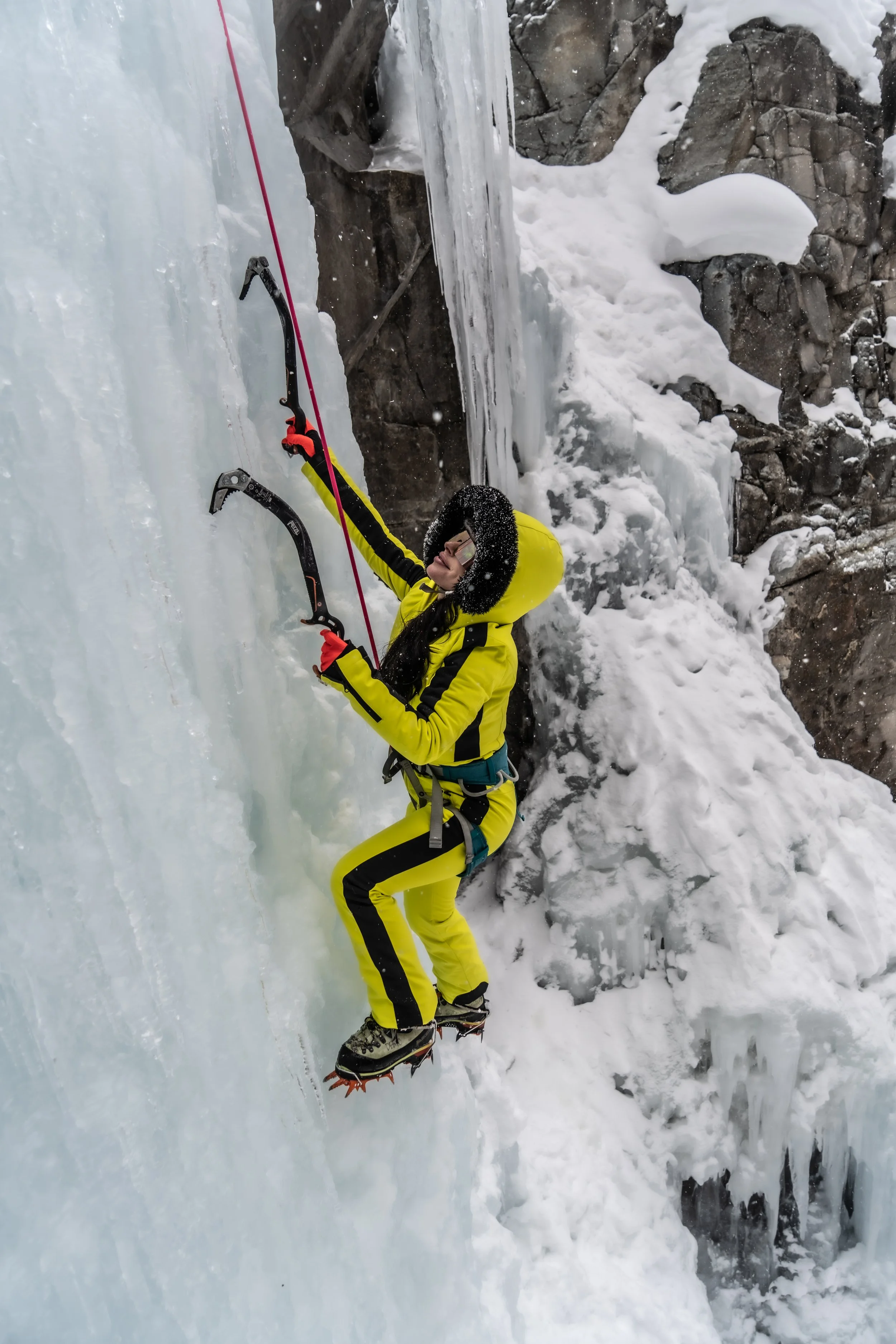 Person ice climbing on a frozen waterfall layered in snow and ice, wearing yellow and black winter gear, an harness, and crampons.