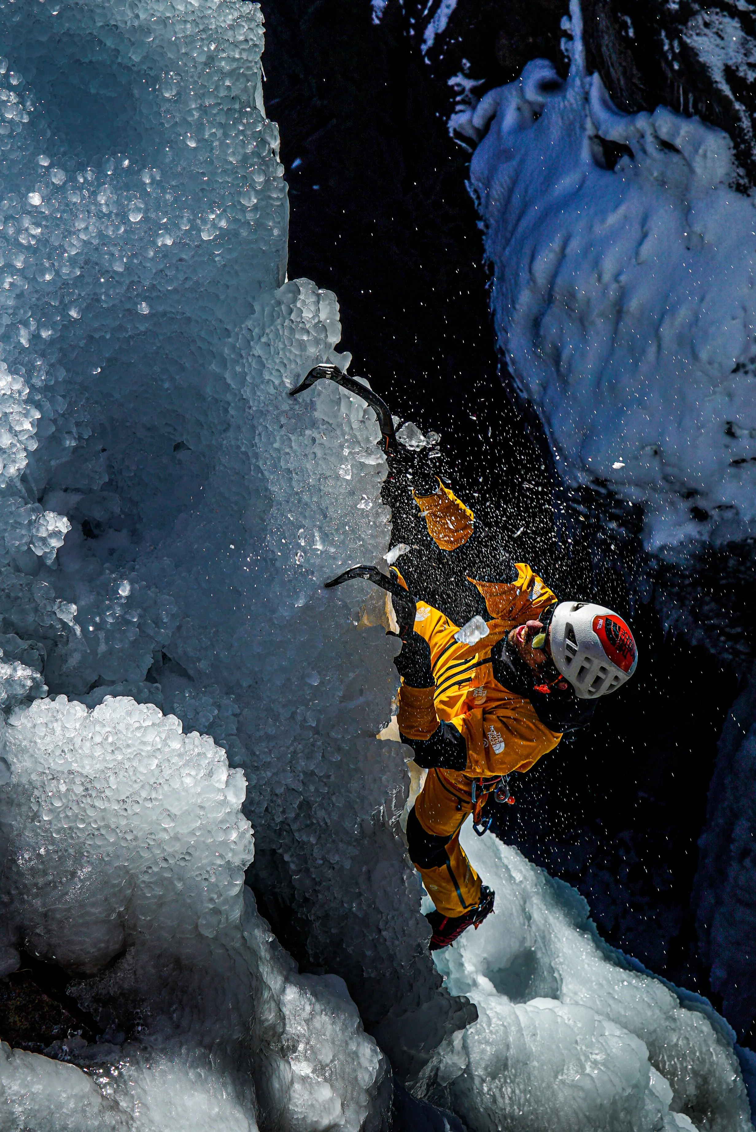 A person in a yellow jacket, wearing a helmet, is ice climbing on a frozen waterfall with ice tools in hand, surrounded by ice and snow.