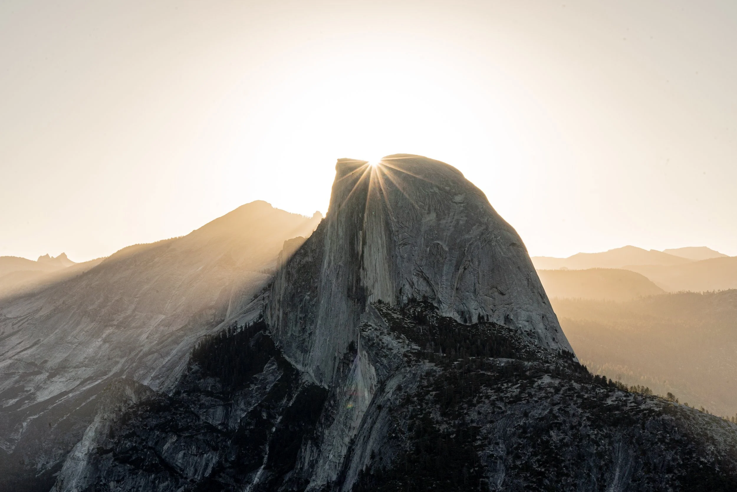 Sun rising behind a prominent mountain peak with surrounding mountain ranges in the distance.