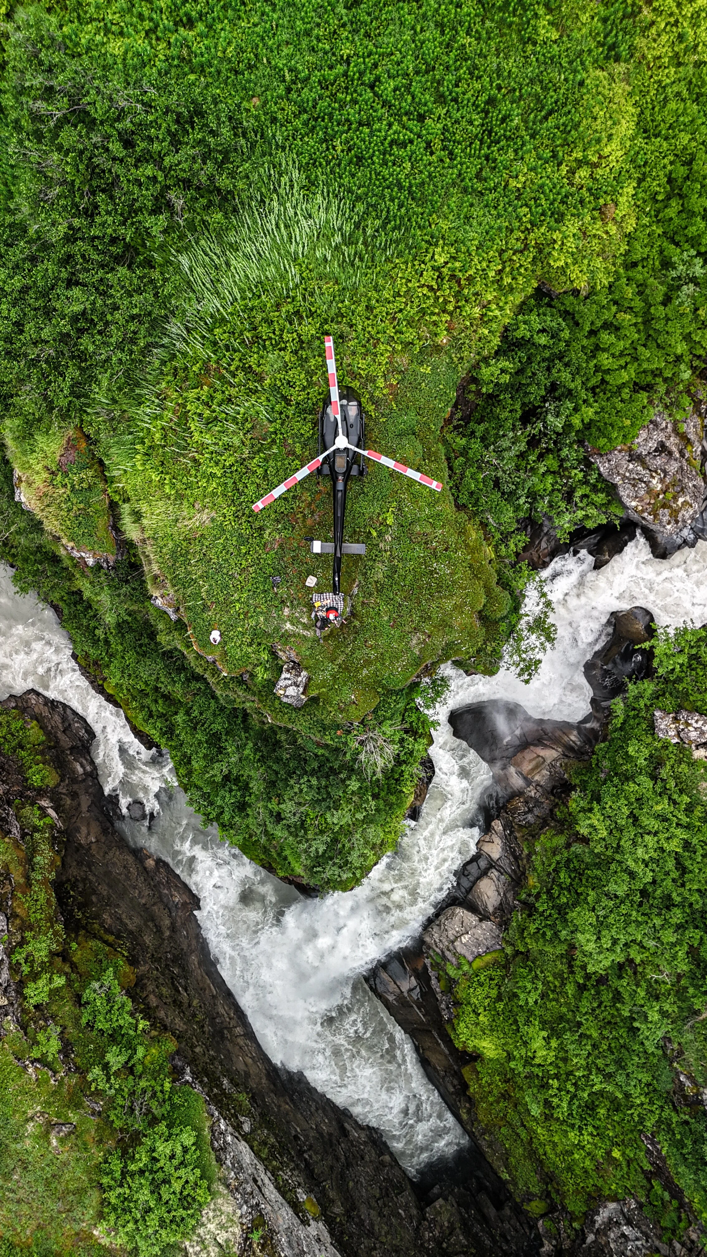 An aerial view of a helicopter hovering over lush green forest and a river with white-water rapids.