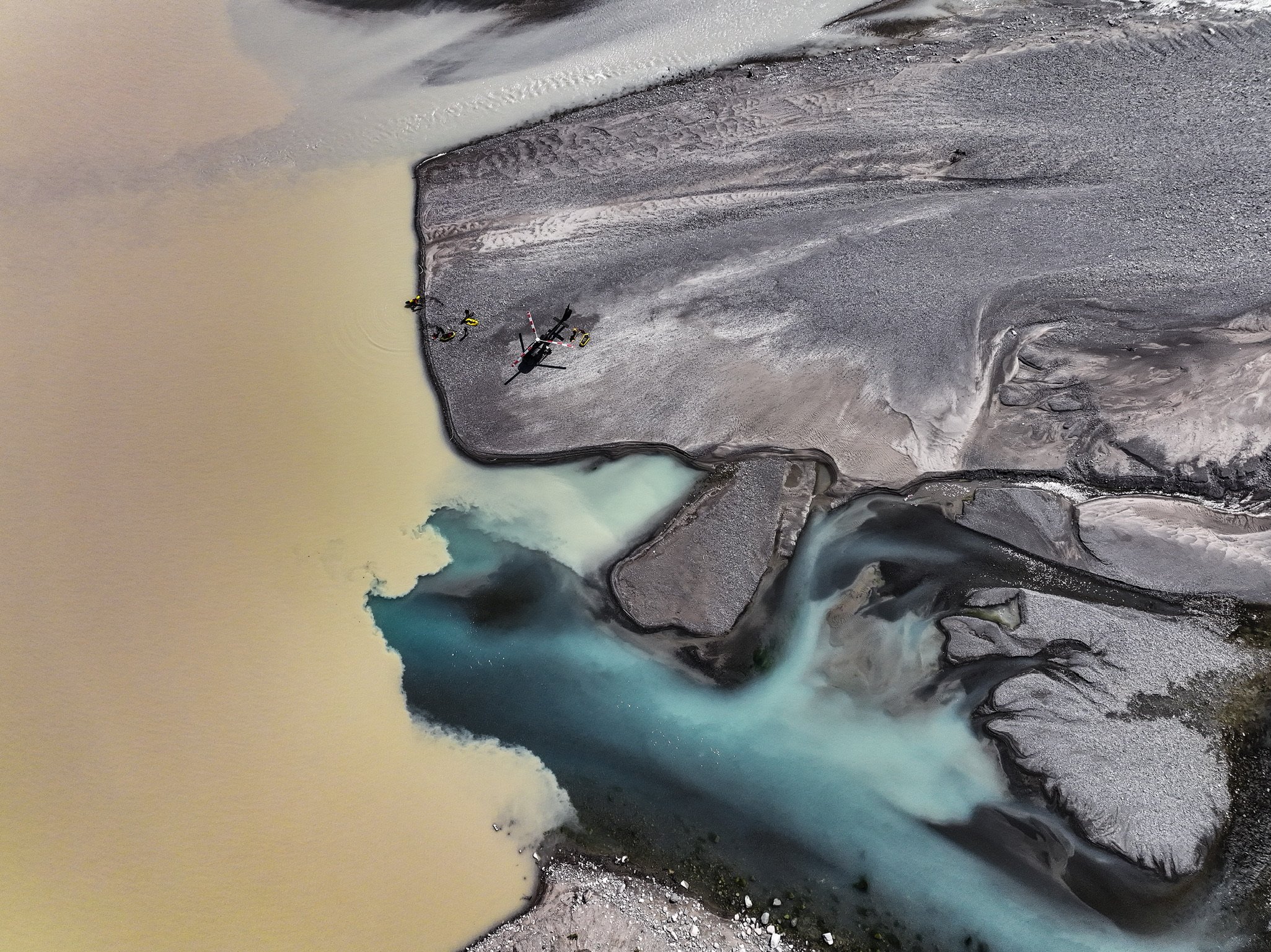 Aerial view of a helicopter and four people near the edge of a flooded area, with a river and landscape showing varying water colors and terrain.