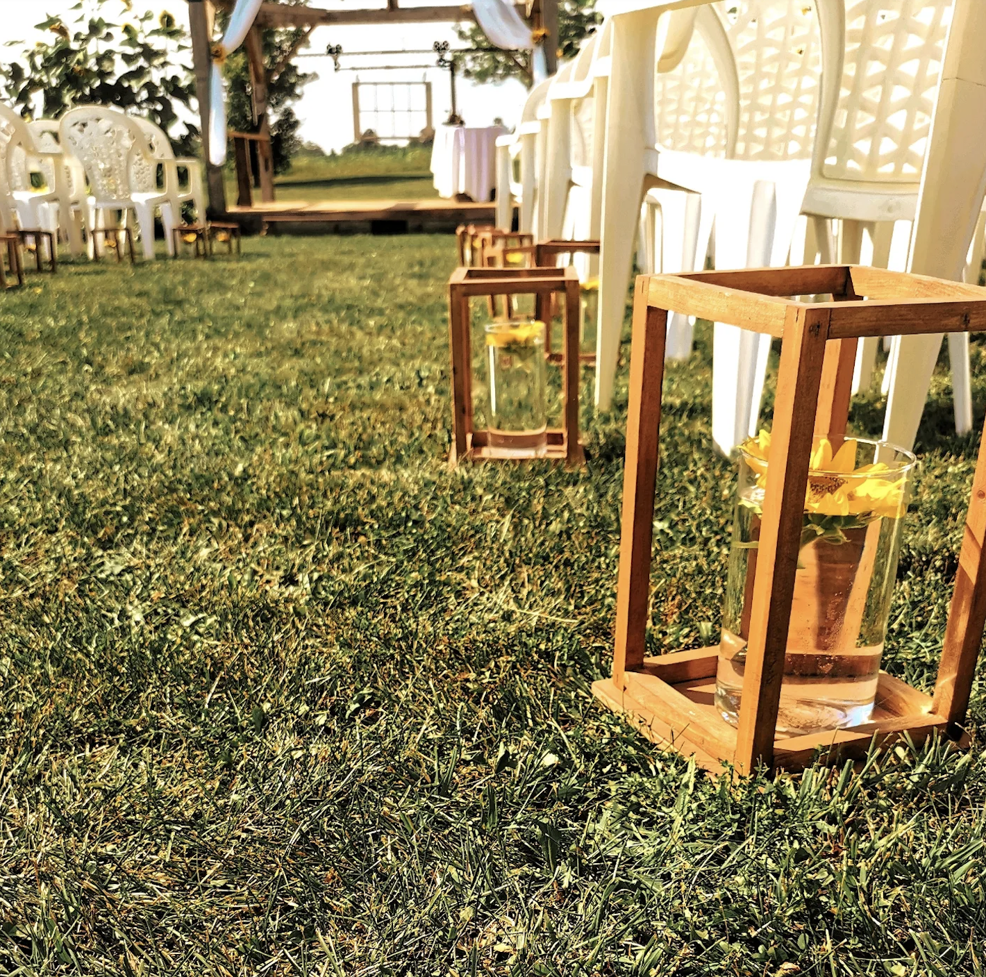 Decorative outdoor setting with white chairs, wooden lanterns with candles, and a small stage in the background, likely for a wedding or event.