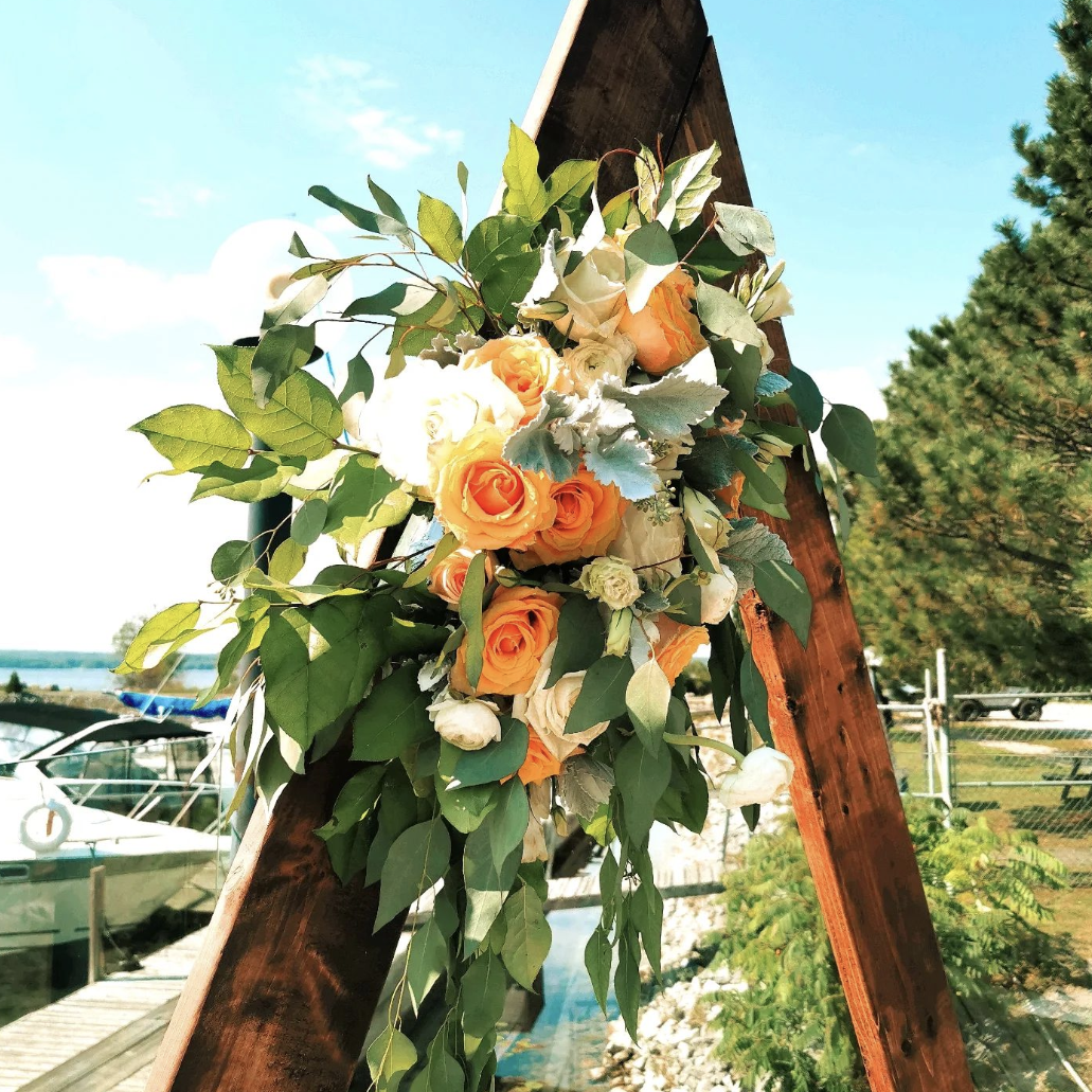 A floral arrangement with orange, white, and cream roses and greenery attached to a wooden A-frame structure, outdoors near water and boats with a clear blue sky.