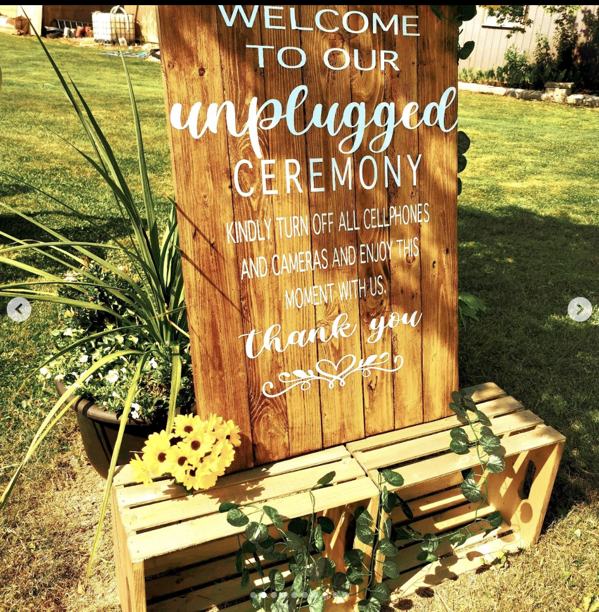 A wooden sign placed on a wooden bench outdoors, with a small potted plant with yellow flowers on the left side and green foliage hanging over the bench. The sign reads: 'Welcome to our unplugged ceremony. Kindly turn off all cellphones and cameras a