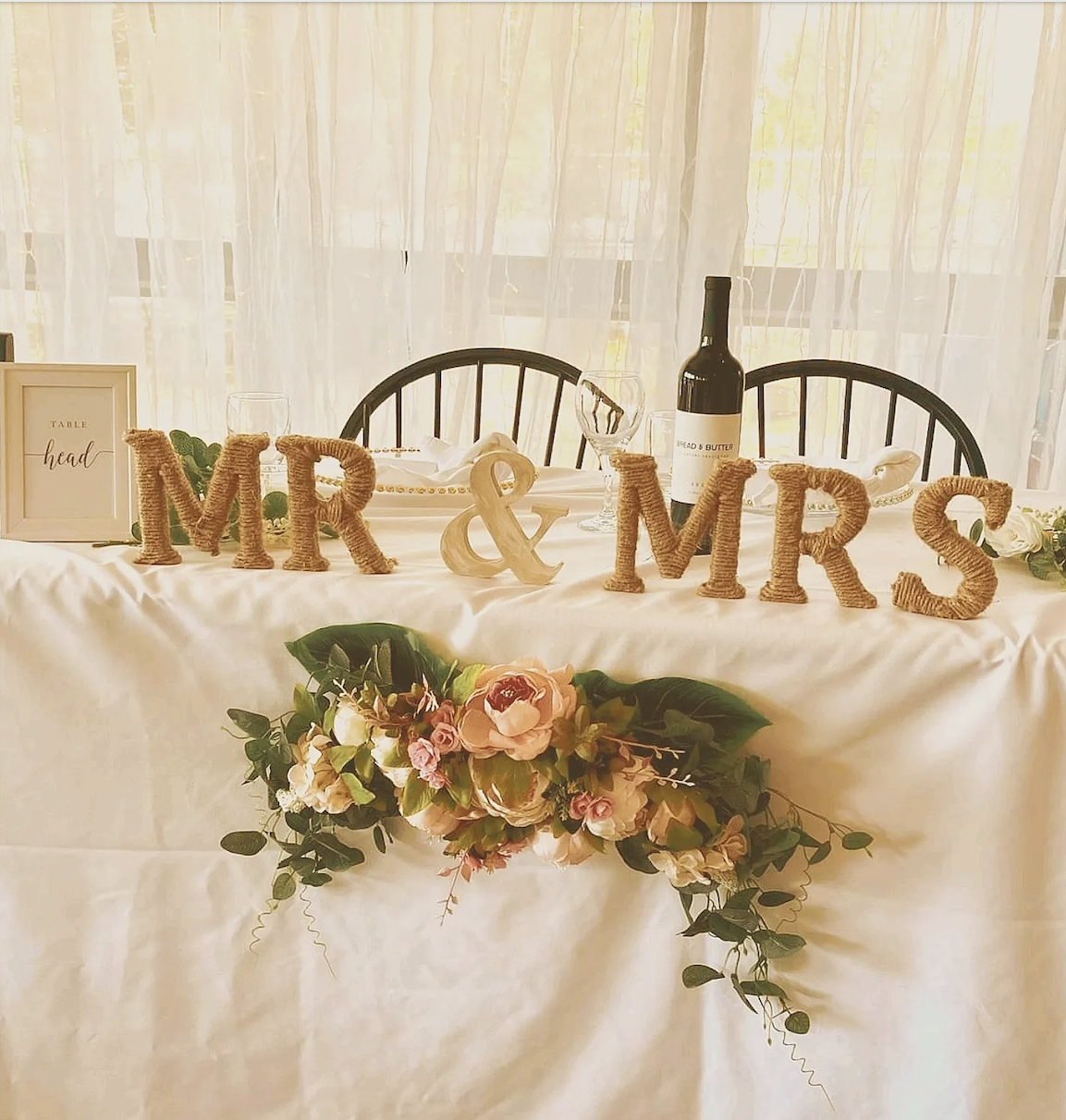Decorated wedding or event table with a sign that reads 'Mr & Mrs', floral arrangements, wine bottle, glassware, and framed sign that says 'Table head'.