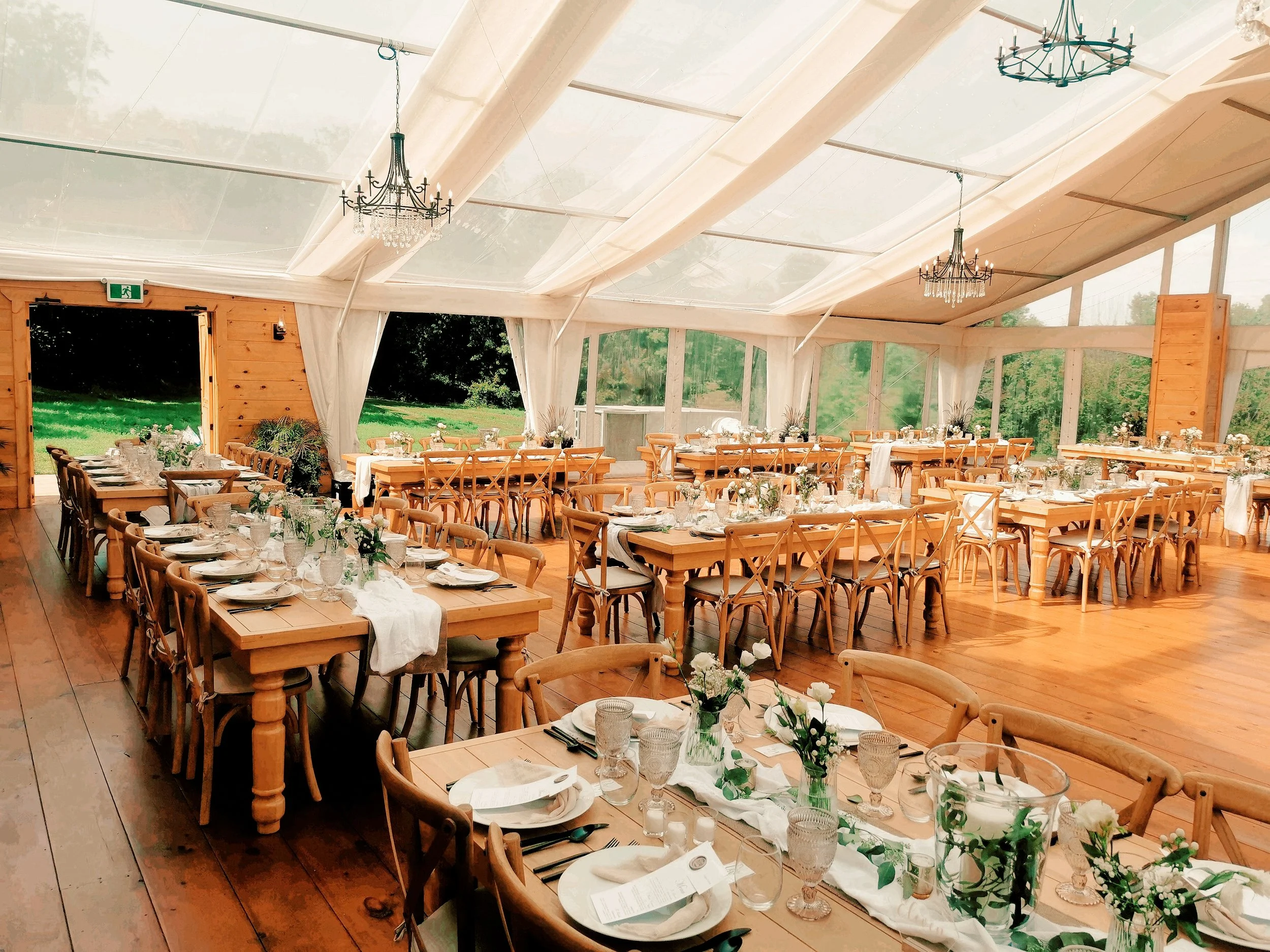 Interior of a large tent with wooden furniture, chandeliers, and tables set for an event, decorated with flowers and tableware, with open sides revealing greenery outside.