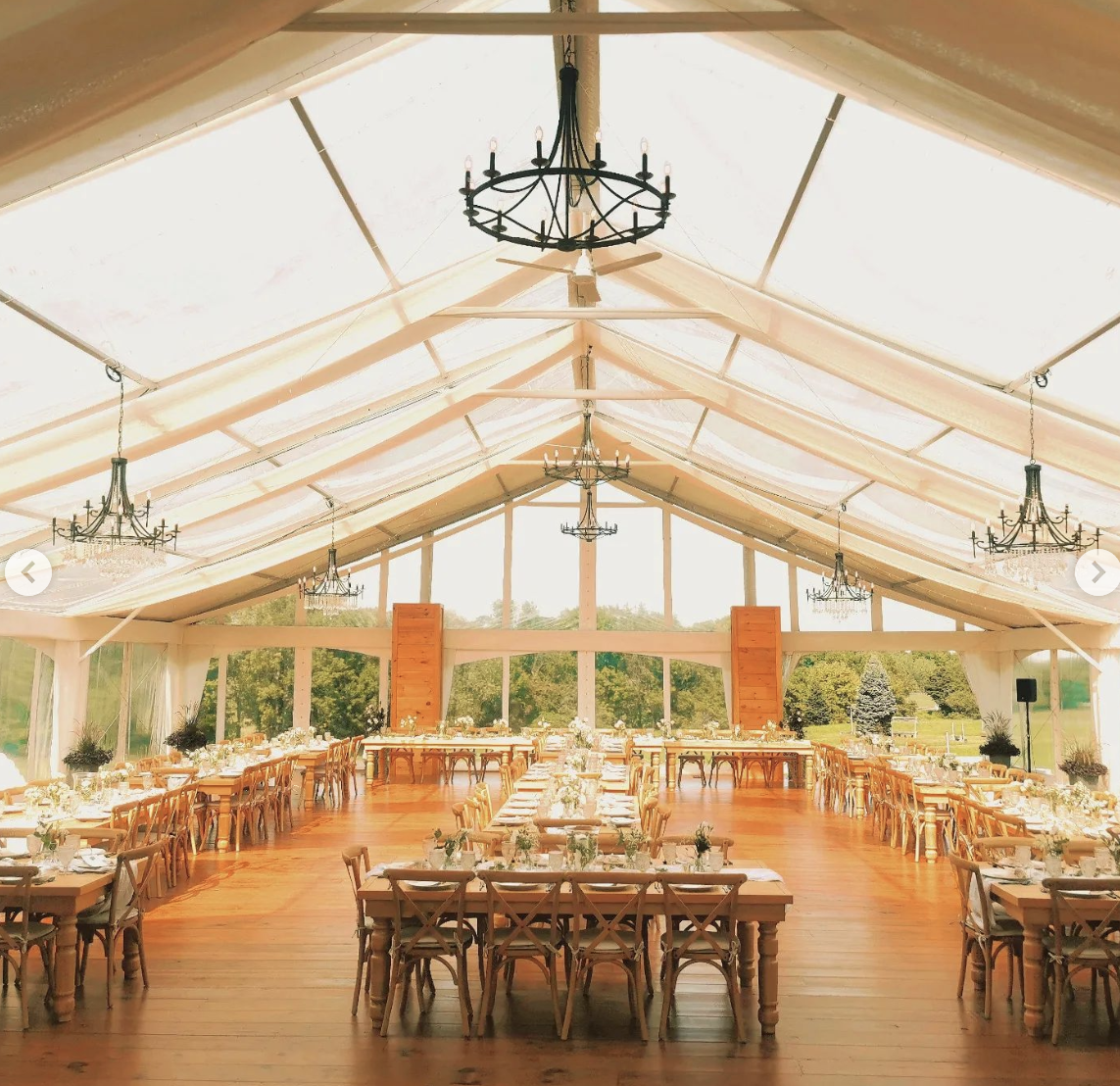 Elegant event tent with wooden floors, decorated tables, chandeliers, and large windows showing a green outdoor landscape.