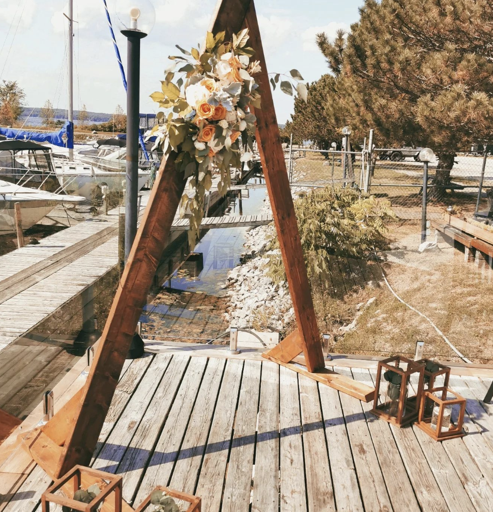 A wooden wedding arch decorated with artificial roses in peach, white, and cream colors, positioned outdoors on a deck near a marina with boats, and surrounded by lanterns and natural scenery.