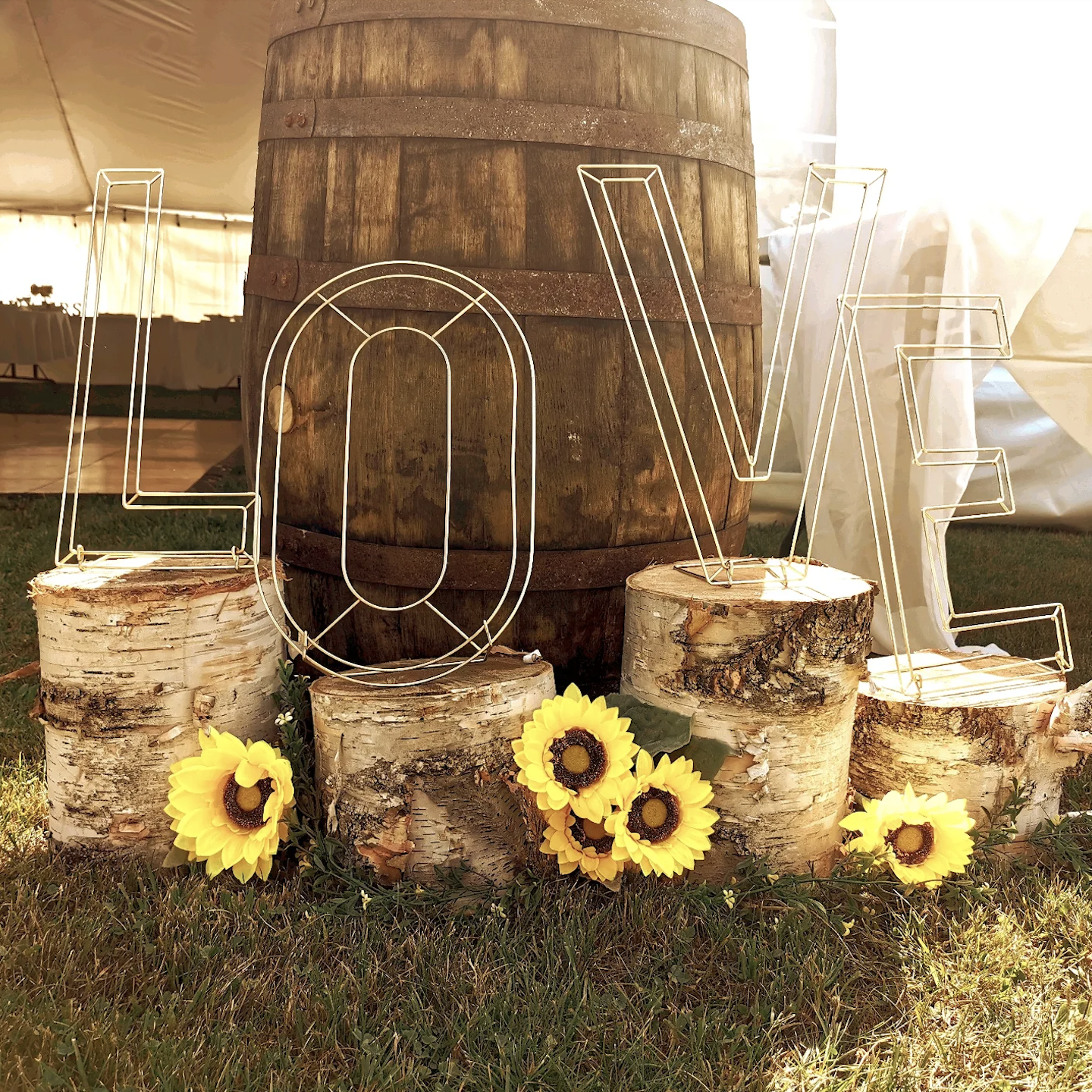 Decorative display with large wooden barrel, log slices, wire letters spelling 'LOVE', and yellow sunflowers on grassy ground under a tent.