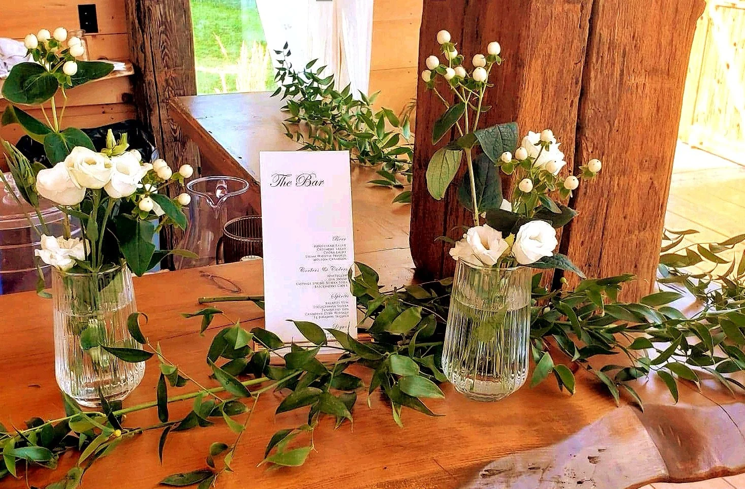 Decorative floral arrangement on a wooden table with glass vases holding white flowers, foliage garland, and a menu card.