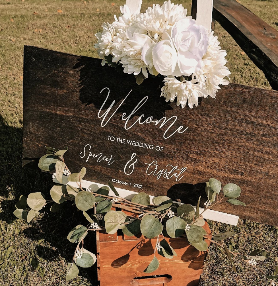 A wooden wedding sign decorated with a white floral arrangement and green eucalyptus leaves, welcoming guests to the wedding of Spence and Crystal on October 1, 2022, outdoors on grass.
