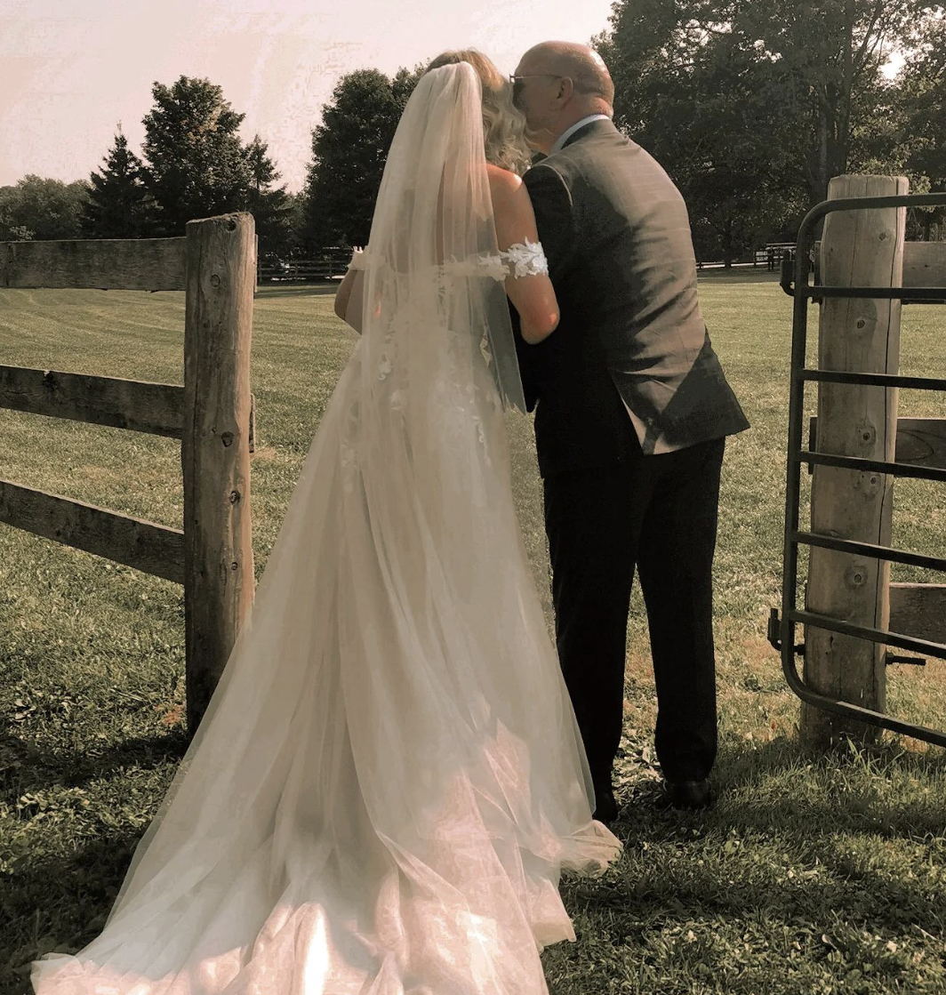 A bride and an older man, possibly her father, sharing a kiss outdoors in a wedding setting during late afternoon or early evening. The bride is wearing a white wedding gown and veil, and the man is dressed in a gray suit. They are standing near a wo