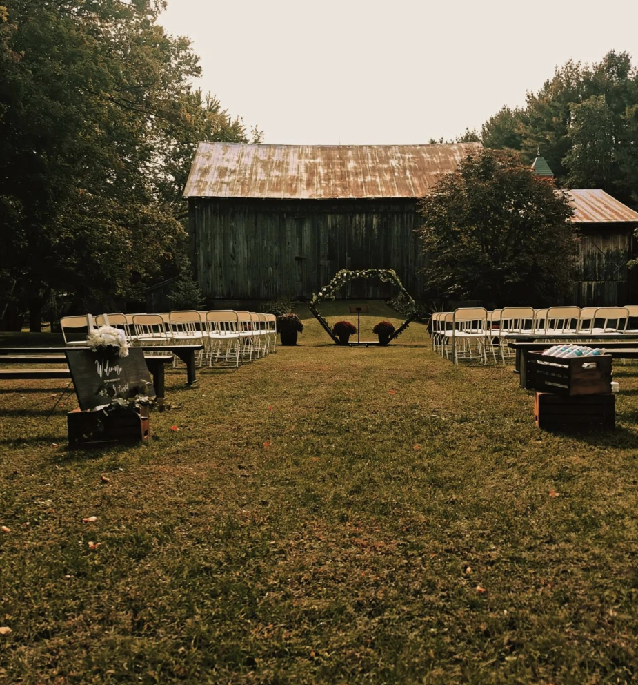 An outdoor wedding setup with white chairs arranged on either side of a grassy aisle, leading to a floral arch at an old barn in the background, surrounded by trees.